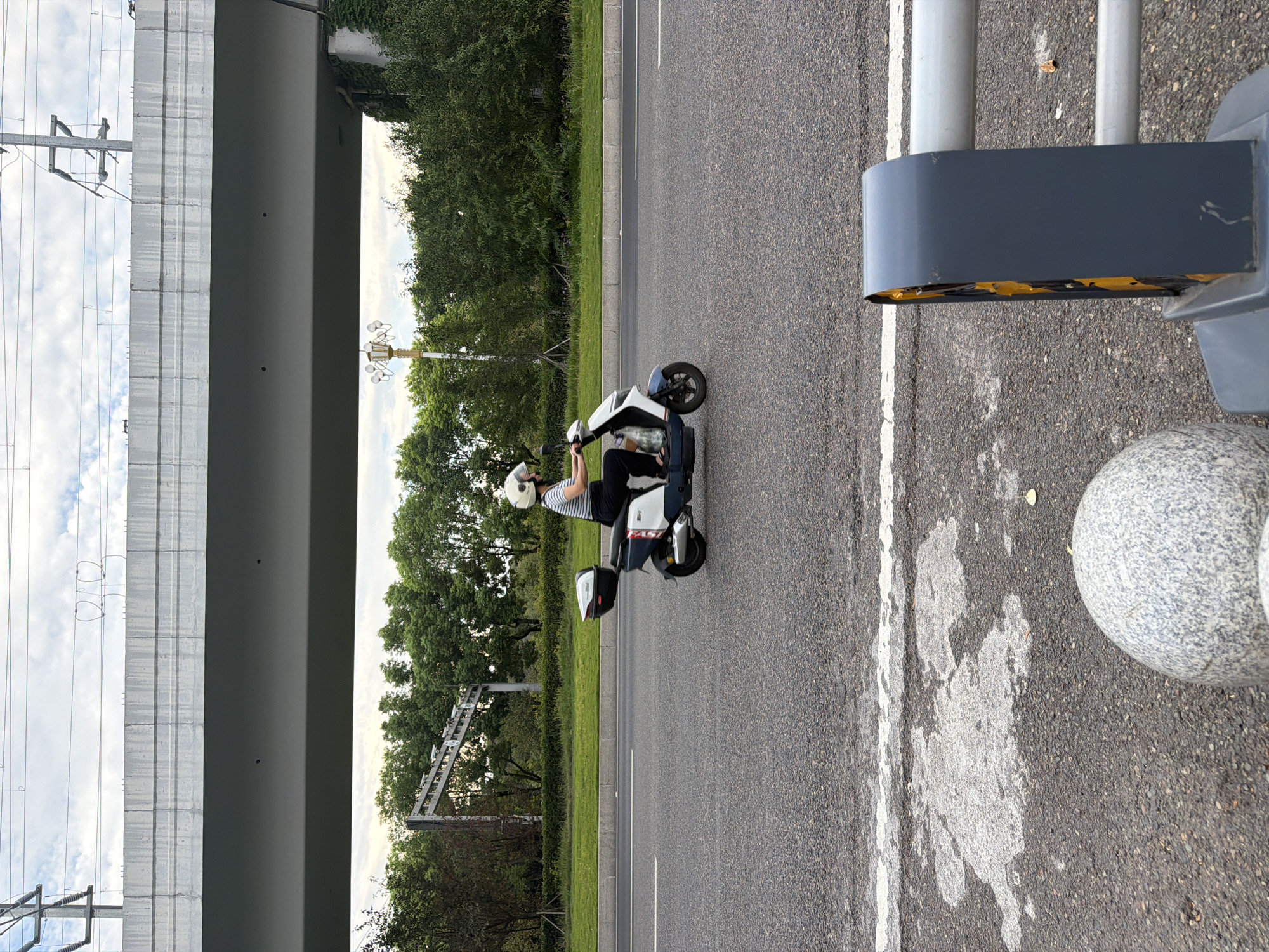 A person wearing a white helmet rides a white and blue electric scooter on a paved road. The scooter has a black storage box on the back. The rider is wearing a striped shirt and dark pants. In the background, there is a large concrete overpass, trees, and a lamppost. The sky is cloudy. The foreground shows part of the road with white markings, a grey spherical bollard, and a grey traffic barrier with yellow reflective material. The scene appears to be in a city.