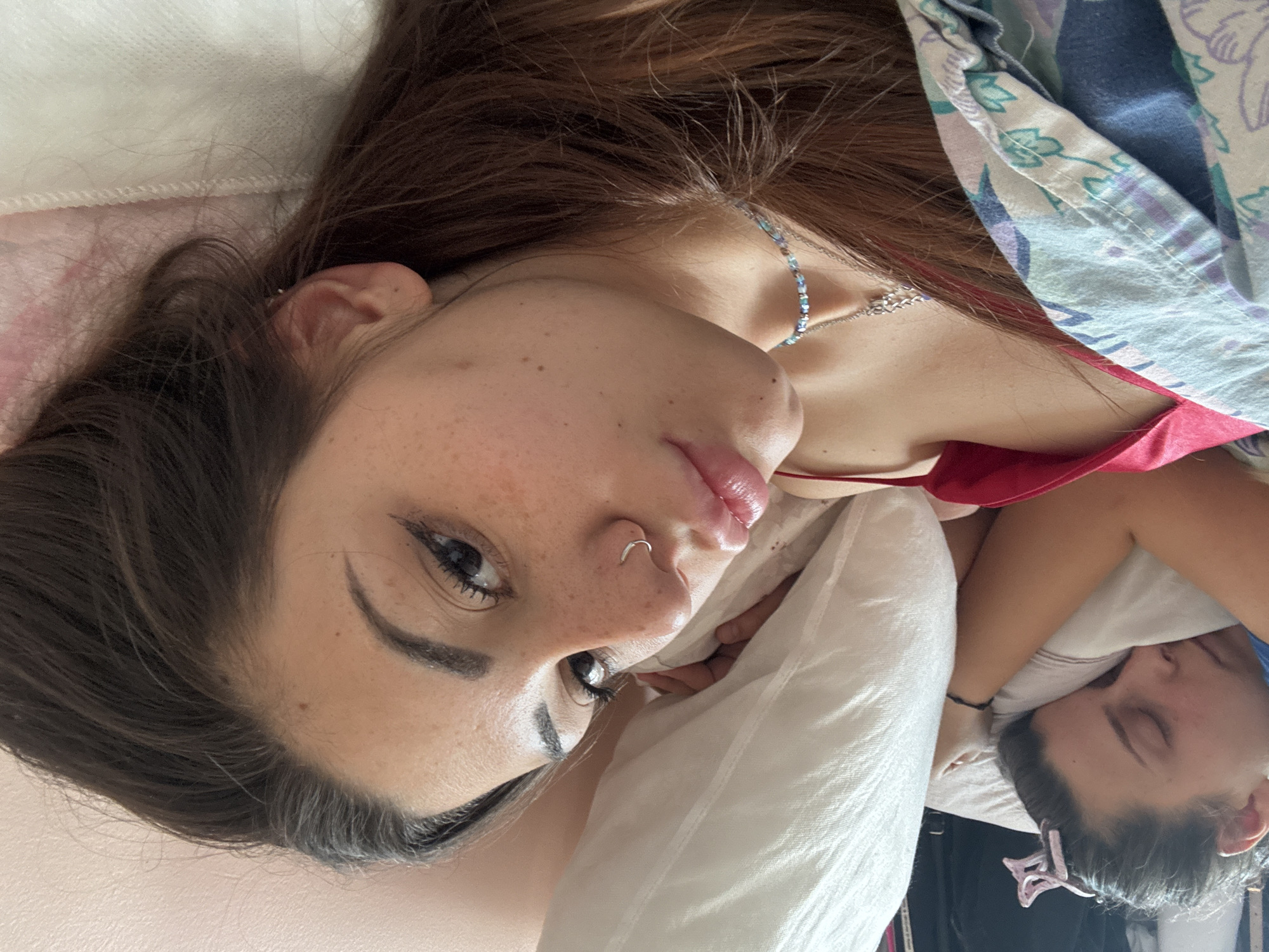 The image, taken in Sofia, Bulgaria, depicts a young woman lying in bed, with her face prominently in the foreground looking towards the camera. Her head is positioned towards the upper-left of the frame, and her long, dark brown hair cascades around her. She has dark eyes enhanced with eyeliner, well-defined eyebrows, and visible freckles across her nose and cheeks. Her lips are slightly parted and appear glossy. A silver hoop nose ring is visible on her left nostril. Around her neck, she wears a delicate, beaded necklace in light blue and silver tones, possibly layered with another thin silver chain. She is dressed in a red top, possibly a tank top or camisole, and is partially covered by a patterned blanket featuring blue, white, and purple colors.
In the lower right background, another individual is visible, sleeping with their eyes closed. This person has dark hair secured by a pink, butterfly-shaped hair clip and wears a dark bracelet on their wrist. Both individuals appear to be resting on white pillows. The scene suggests a relaxed indoor setting, likely a bedroom. The main subject's expression is neutral and calm, while the background individual is clearly asleep. There is no visible text in the image.