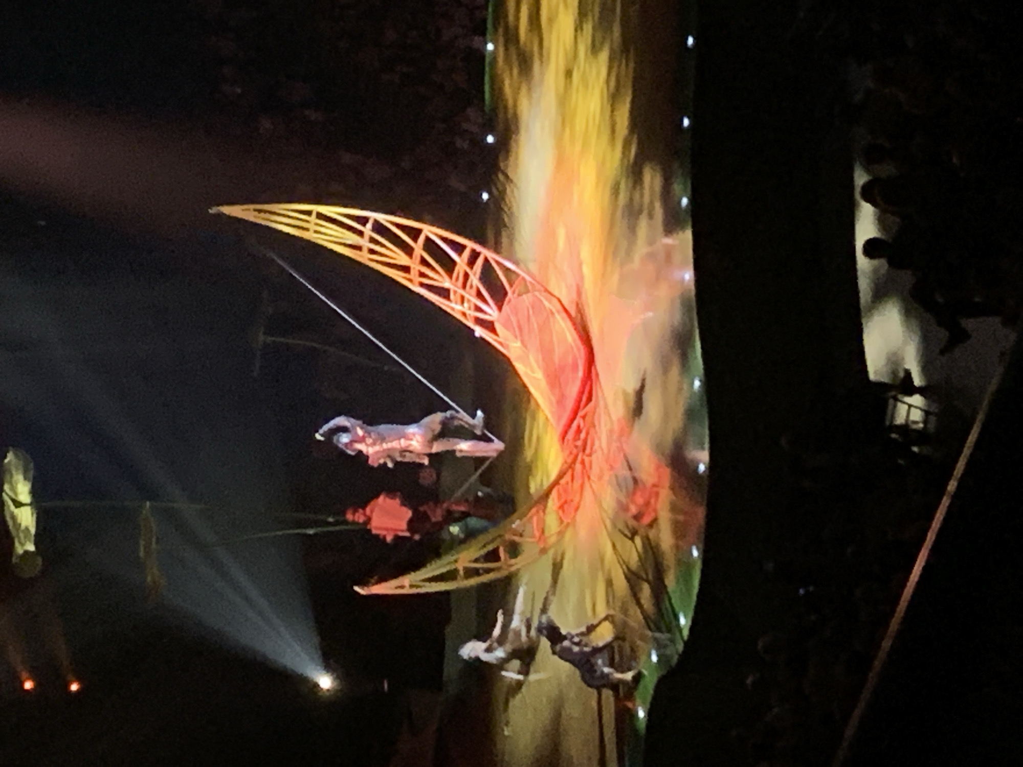 The image captures a Cirque du Soleil performance, possibly in Belgrade, Serbia, given the context. A large, crescent-shaped structure with an orange frame is suspended mid-air, with performers attached to it in various positions. A large screen behind the structure displays vibrant colors, primarily yellow and orange, suggesting flames or a fiery backdrop. To the left, a spotlight illuminates another performer suspended by ropes. On the right, partially obscured by darkness, are audience members. The overall scene conveys a sense of spectacle, with aerial acrobatics and dramatic lighting.