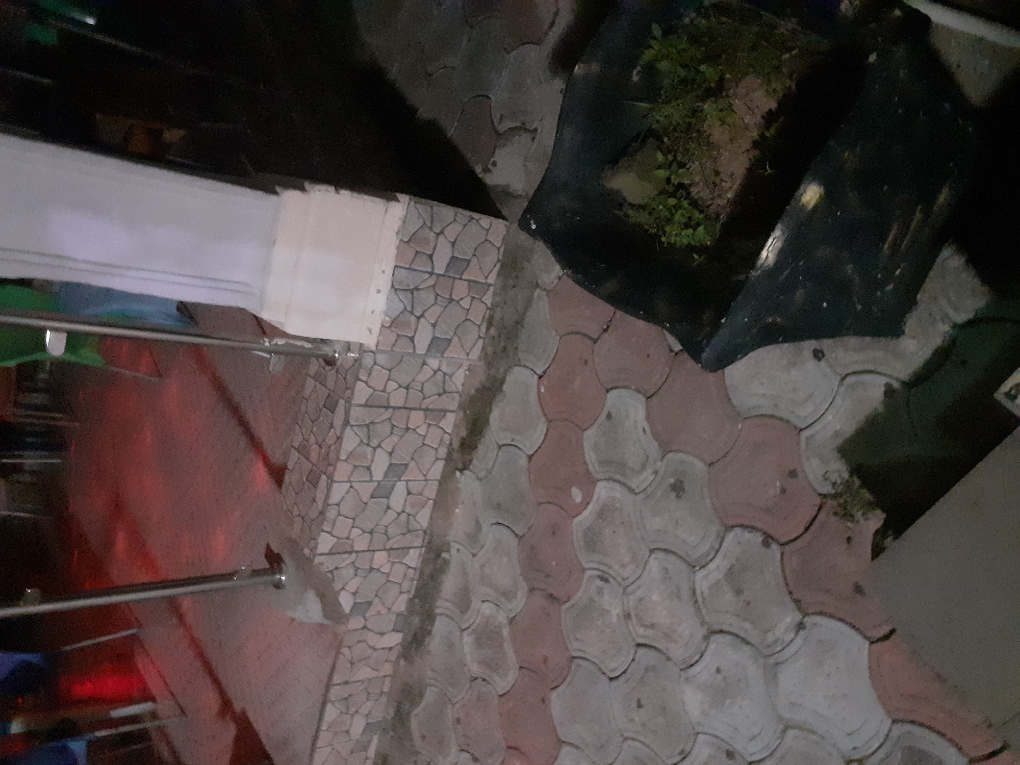The image depicts an outdoor scene in Jalingo, Nigeria, likely at night or during low light conditions, given the artificial lighting casting red hues on the ground. In the foreground, a black plastic planter box contains soil and some green vegetation, suggesting a neglected or underdeveloped planting area. The ground is paved with interlocking concrete blocks, featuring a pattern of alternating grey and reddish-brown sections. To the left, a metal railing is visible, anchored to a concrete base. Behind the railing, a tiled step leads upwards, with the tiles having a mosaic-like appearance of small, irregular stone pieces in shades of grey and beige. A prominent white pillar stands to the right, its base integrated with the tiled step. The background is dark, with indistinct shapes of furniture or structures and the glow of red lights, hinting at a seating or recreational area. The overall impression is that of an outdoor public space, possibly a garden, patio, or restaurant seating area, in a state of disrepair or undergoing development.