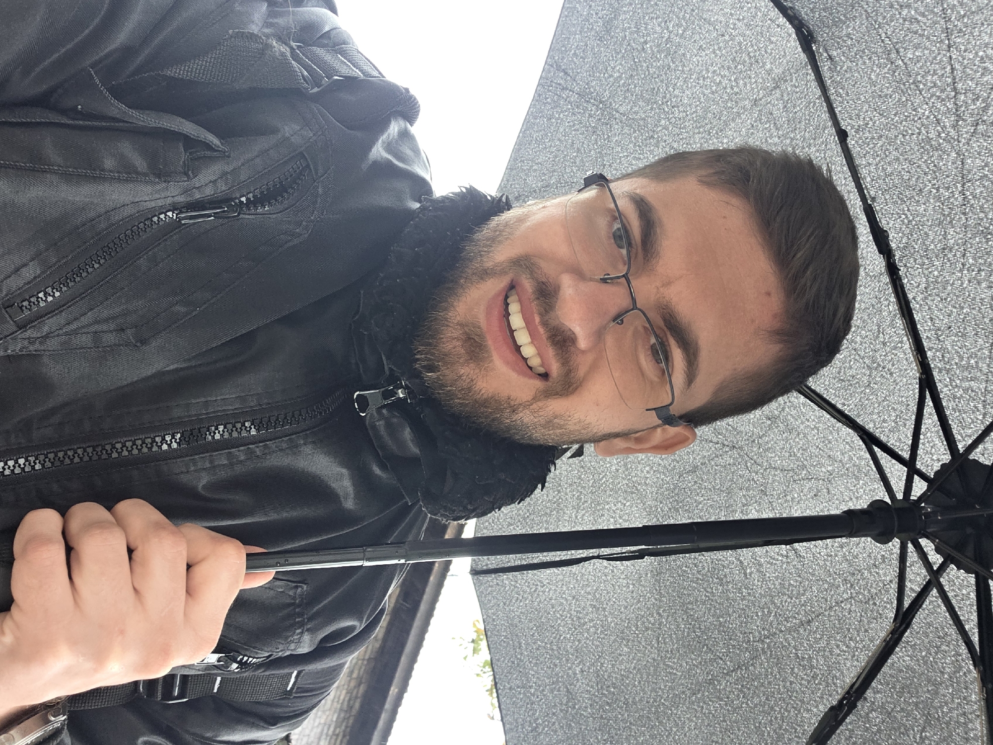 A man, appearing to be in his 20s or 30s, is featured in a close-up, vertically oriented photograph. He has short dark hair, a beard, and wears dark-rimmed glasses. A broad, cheerful smile reveals his teeth, suggesting a happy or amused mood. He is dressed in a black jacket featuring prominent zippers and a dark, possibly faux fur, collar. His right hand is visible in the lower left, holding the black handle of an open umbrella. The umbrella dominates the right half of the image, showcasing its textured, light grey or silver underside and black structural ribs. The background is brightly lit, likely an overcast sky, with faint glimpses of a building and green foliage in the distance, indicating an outdoor, possibly urban, environment. The use of an umbrella suggests rainy or highly reflective conditions. No visible text is present. The image's orientation is rotated 90 degrees counter-clockwise from a standard portrait view. The scene is located in Plovdiv, Bulgaria.