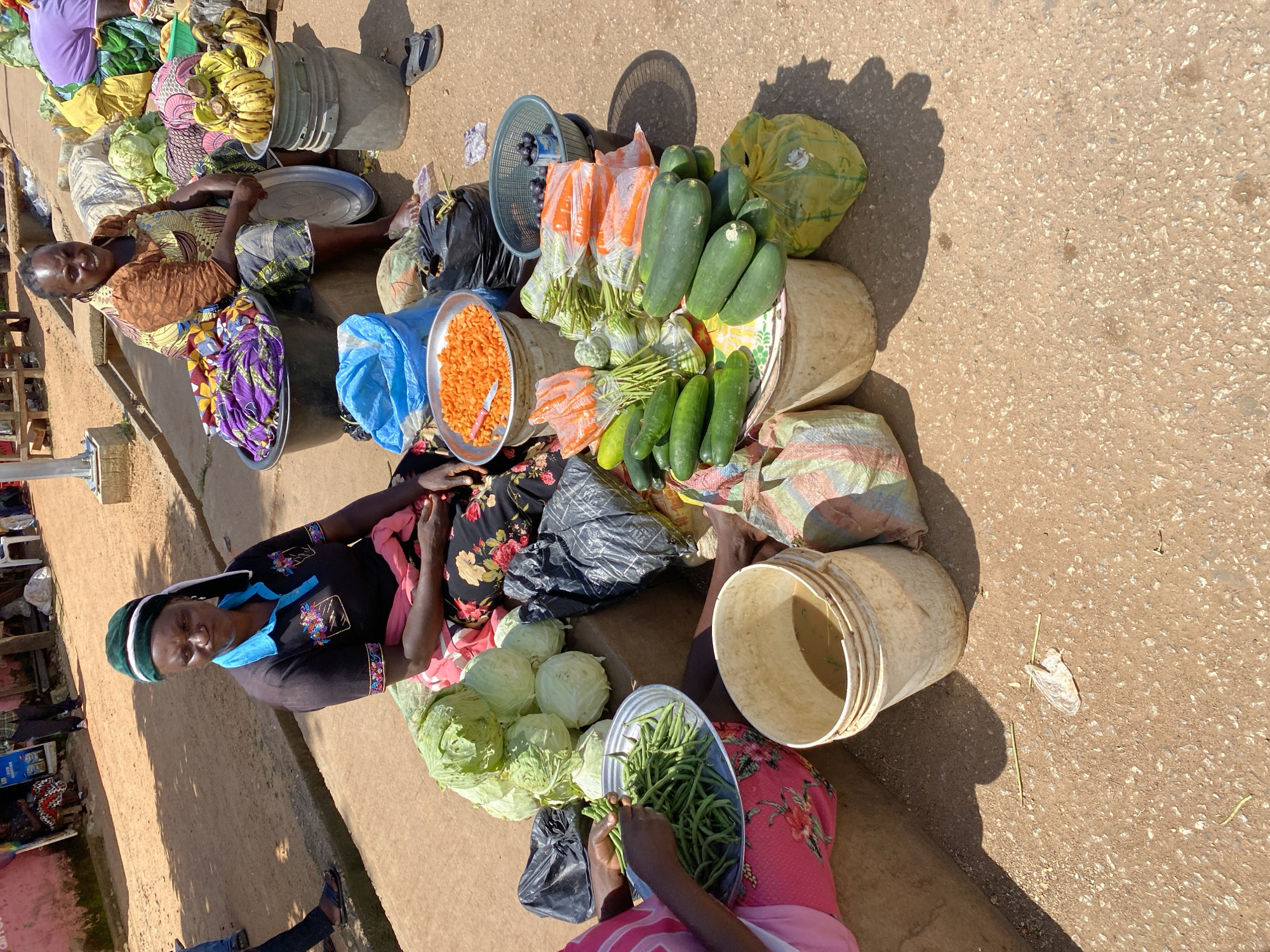At an outdoor market, likely in Bukuru, Nigeria, two women are visible amidst piles of fresh produce. In the foreground, a woman wearing a black t-shirt with colorful floral embroidery and a green headwrap sits behind a selection of vegetables. She is handling a pile of green beans, which are arranged in a metal tray. To her right, a large, off-white plastic bucket contains murky water. Behind her, a mound of cabbages is visible. To the right of the cabbages, a second woman, wearing a patterned dress and apron, is seated. She is surrounded by produce, including bananas on a silver tray atop a grey bucket, and several bunches of carrots wrapped in plastic. Other vegetables, such as cucumbers, are also displayed. In the background, more produce, including large bags of yellow and purple items, are piled high, suggesting a bustling market environment. The ground is a mix of red earth and concrete.