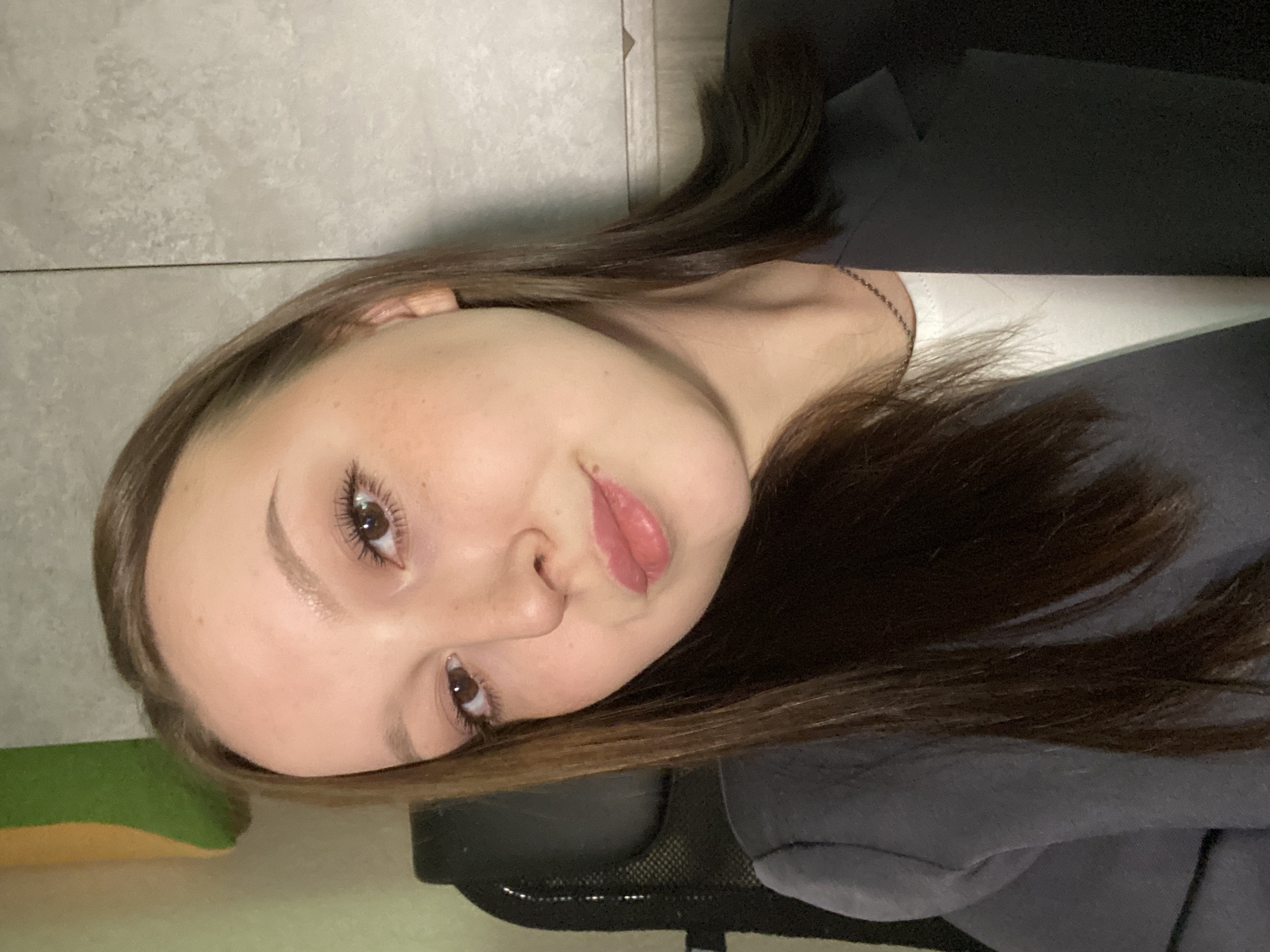 A portrait of a young woman with light skin, freckles, and brown hair. She has brown eyes and slightly parted lips. She wears a white top and a dark-colored jacket or sweatshirt. The background is a combination of a light-colored wall and darker objects, including what appears to be a black chair. The image is tilted to the side. There is no information to indicate the location is the city of Chaikovsky, Russia.