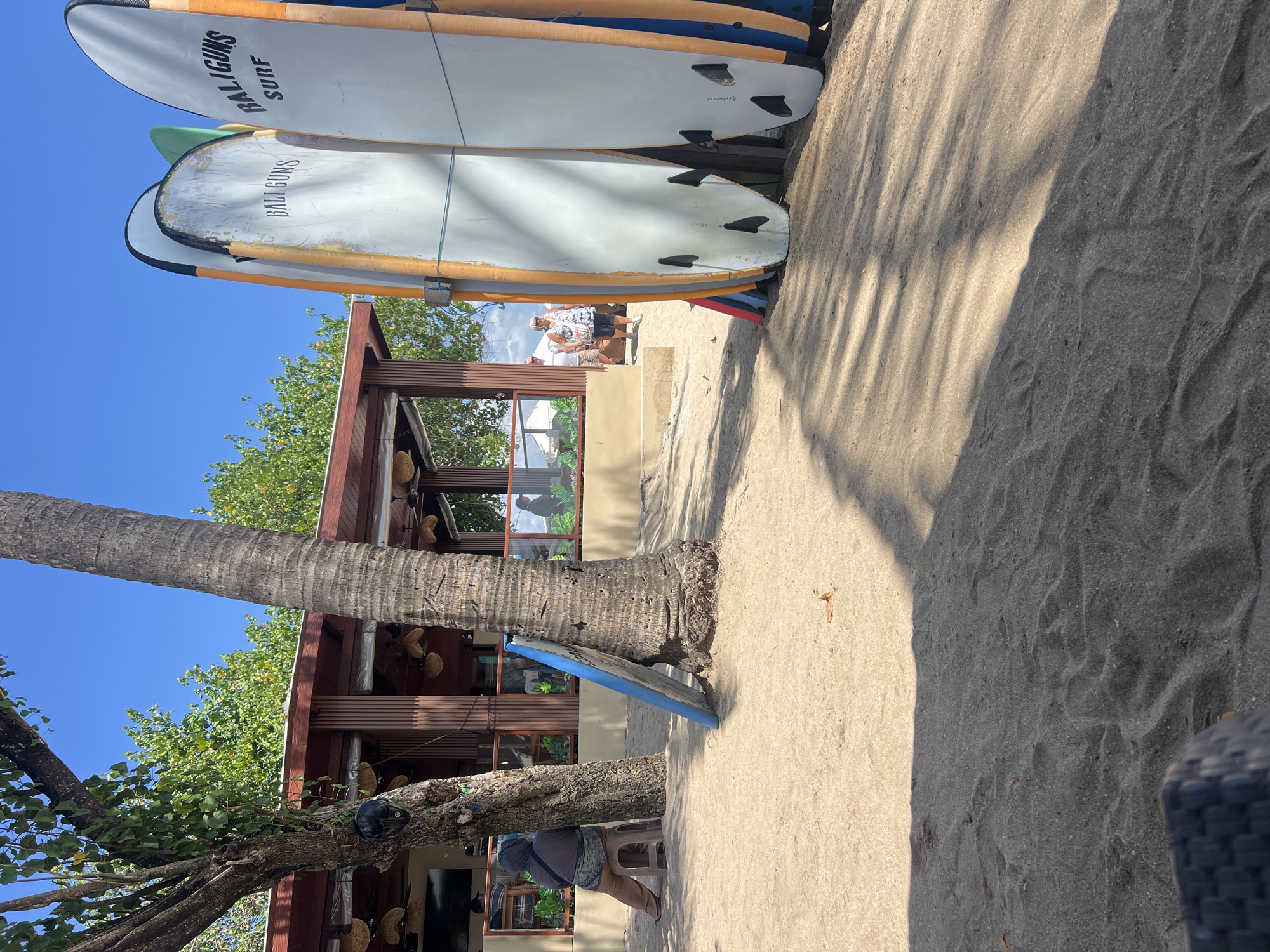 A beach scene in Kuta, Indonesia features a row of surfboards propped against a fence. The surfboards are mostly white with orange and blue accents. The text "BALIGUNS SURF" is visible on at least two of the boards. In the background, a wooden building with a thatched roof serves as a cafe or restaurant, with patrons visible through the windows. Trees with lush green foliage provide shade, and a person is seated on a stool near one of the trees. The ground is covered in sand, with shadows cast by the trees and surfboards. The sky is clear and blue.