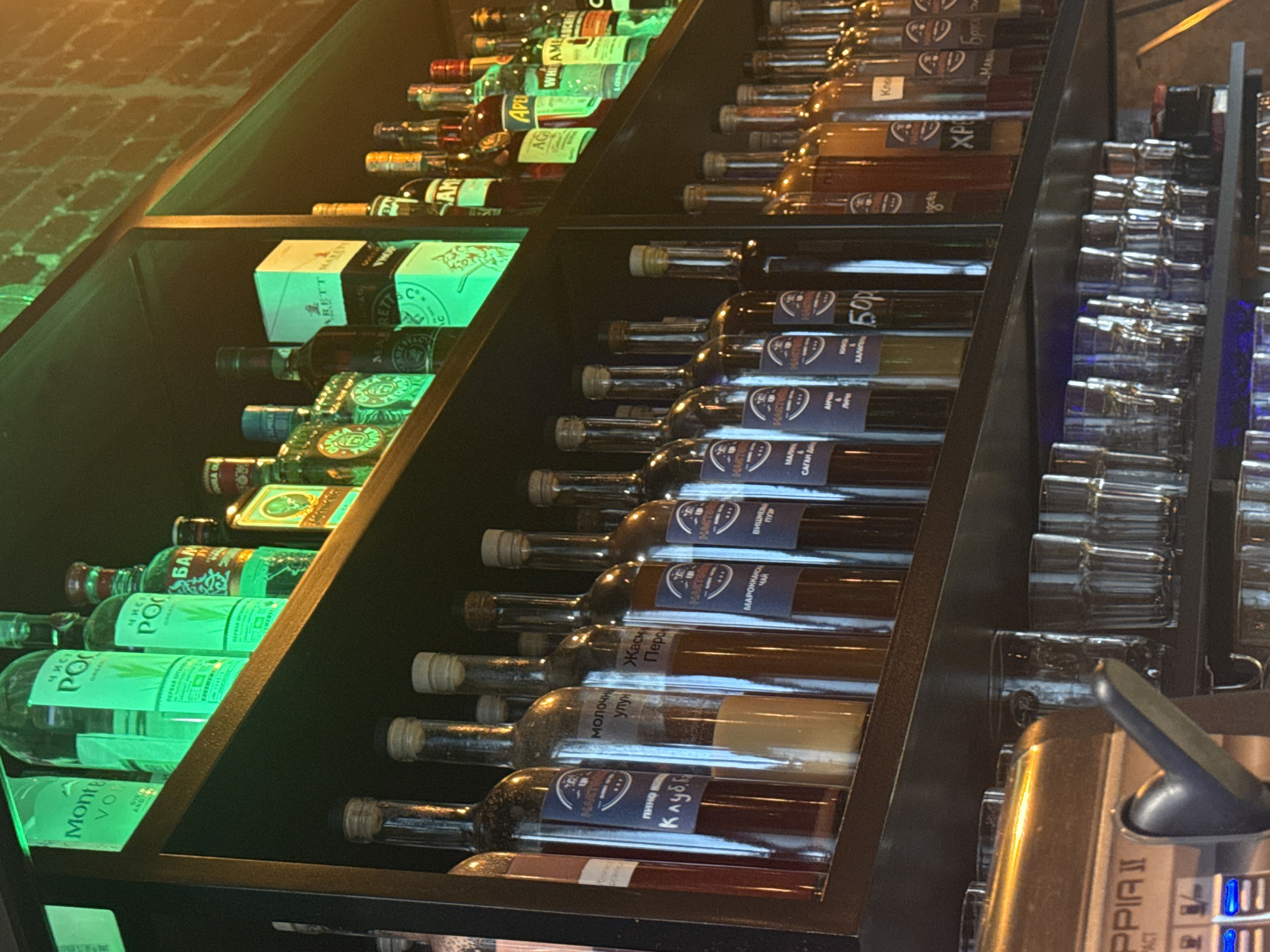 A well-stocked bar shelf presents a variety of alcoholic beverages. The shelf on the left features brands such as "Monte VO" and "Чист PO", suggesting a selection of vodkas or similar spirits. The middle shelf holds "Amaretti Marti" and other unidentified bottles, while the right shelf contains an array of clear drinking glasses. Some bottles bear labels in Cyrillic script, such as "Молочный улун" and "Жасм Перо", hinting at local production or flavors. A dispenser labeled "PPIA II" stands at the edge of the shelf. The scene could be located in a bar or restaurant in Moscow, Russia, given the presence of Russian writing.
