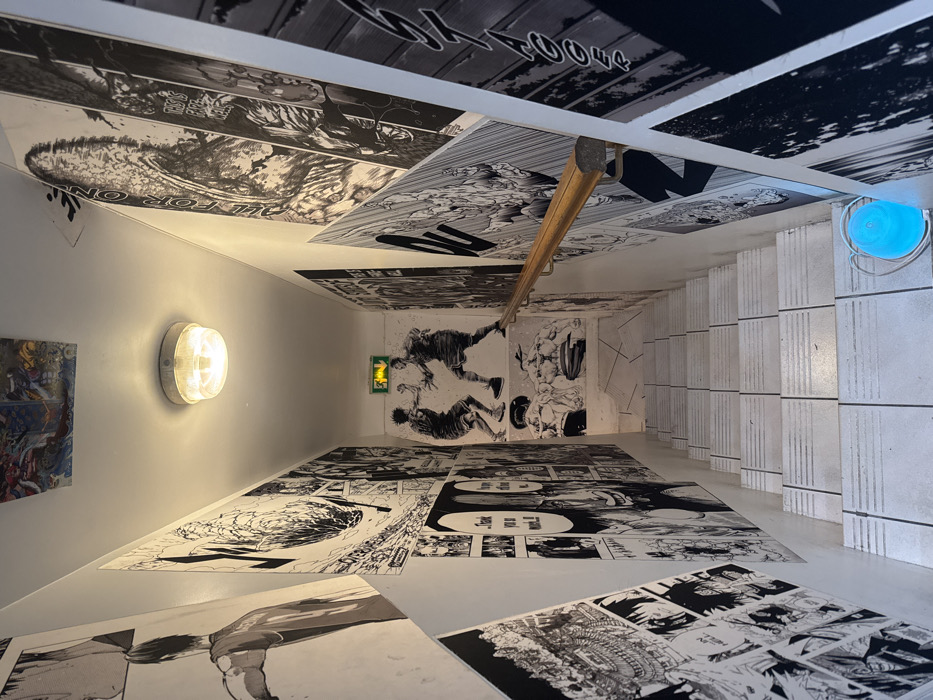 The image depicts a downward-sloping, well-lit interior staircase within a building in Paris, France. The walls along the stairwell are entirely covered with large, black-and-white prints of various manga panels and illustrations. These prints feature dynamic action scenes, character portraits, and speech bubbles. A prominent illustration on the far wall at the landing portrays two athletic figures in an aggressive stance, possibly engaged in boxing or martial arts, with another illustration of two muscular, bare-chested figures beneath them. An emergency exit sign, green with a white running pictogram, is visible on the upper section of the far wall. The staircase itself is constructed from white tiles with horizontal grey grout lines defining each step. A light-toned wooden handrail with metallic brackets extends along the right wall from the top of the stairs downwards. A single circular, translucent ceiling light fixture provides illumination from above. A small, light blue object, resembling a container or bag, is present on the tiled floor at the bottom of the stairs to the right. No individuals are present in the scene.