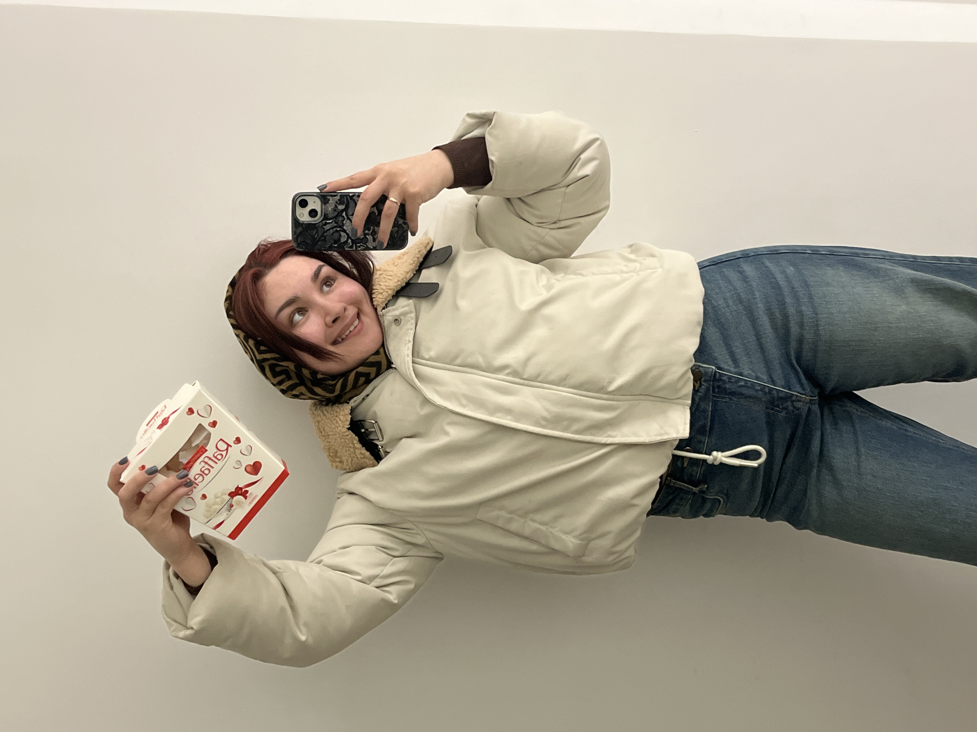 In the city of Neftekamsk, Russia, a young adult female stands indoors against a plain, light-colored wall, with a vertical wall junction visible on the right. She is seen from the waist up, dressed in a light beige puffy winter jacket featuring a brown faux-shearling collar and visible front pockets, a dark brown long-sleeved shirt, and blue denim jeans with a white drawstring detail on the left side. She wears a patterned headscarf with brown and yellow/beige geometric designs covering her head, allowing reddish-brown hair to frame her face. Her fingernails are painted a dark blue-gray.
With her right hand raised, she holds a white, partially open box of "Raffaello" chocolates, decorated with red hearts and inverted "Raffaello" text, with white confectionery visible inside. Her left hand holds a dark-colored smartphone, possibly an iPhone, encased in a black textured cover, positioned for a selfie. A gold-colored ring is visible on her left ring finger. The individual is smiling broadly, with her gaze directed towards the phone.