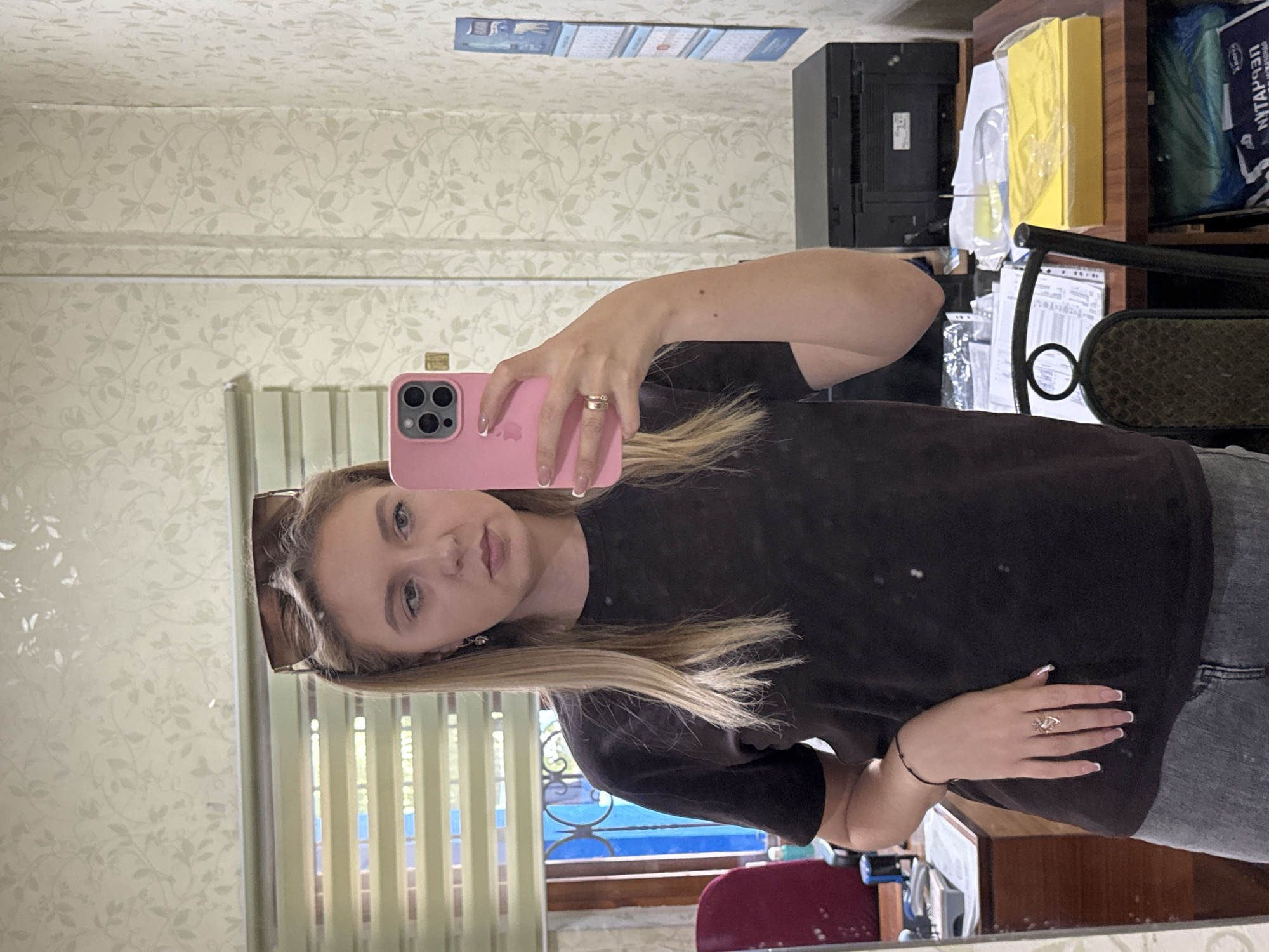 A young woman with blonde hair is taking a mirror selfie in an indoor setting. She is wearing a dark t-shirt and jeans. She has sunglasses perched on her head, and is holding a pink smartphone in her right hand. There are rings on her fingers, and a bracelet on her left wrist. The background shows a wall with patterned wallpaper, a window with blinds, and a desk with a printer and papers.