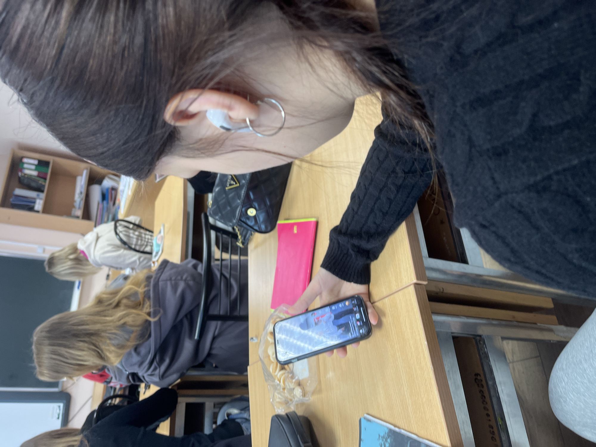 The image captures an indoor scene, likely a classroom or lecture hall, with students seated at wooden desks. The perspective is from behind and slightly above a person in the foreground, focusing on their ear, hair, and hand holding a smartphone.
The person in the foreground has long dark hair and wears a dark, possibly knitted or ribbed, long-sleeved top. They have a white earbud in their right ear and a silver hoop earring. Their right hand holds a black smartphone, which displays a video or live stream featuring a person with dark hair in a light-colored top, speaking or performing. The phone screen also shows typical video player interface elements. Next to the phone on the wooden desk is an open plastic bag containing light-colored snacks, possibly small cookies or crackers, and a bright pink notebook or folder. A black quilted bag with a prominent "GUESS" logo is visible on the chair or desk to the left of the foreground person.
In the background, two other individuals are visible, also seated at desks. One person has long blonde hair and is wearing a grey hoodie, while another further back has lighter hair and is wearing a light-colored top. Both appear to be engaged in an activity at their desks. The room features a wooden bookshelf filled with books and binders, and parts of a white board or screen are visible on the left side of the frame. The desks are simple, made of light-colored wood with metal frames. One desk frame in the bottom right corner shows a serial number "0621000200". The general lighting suggests it is daytime. No specific emotions are discernible from the individuals' obscured faces.
