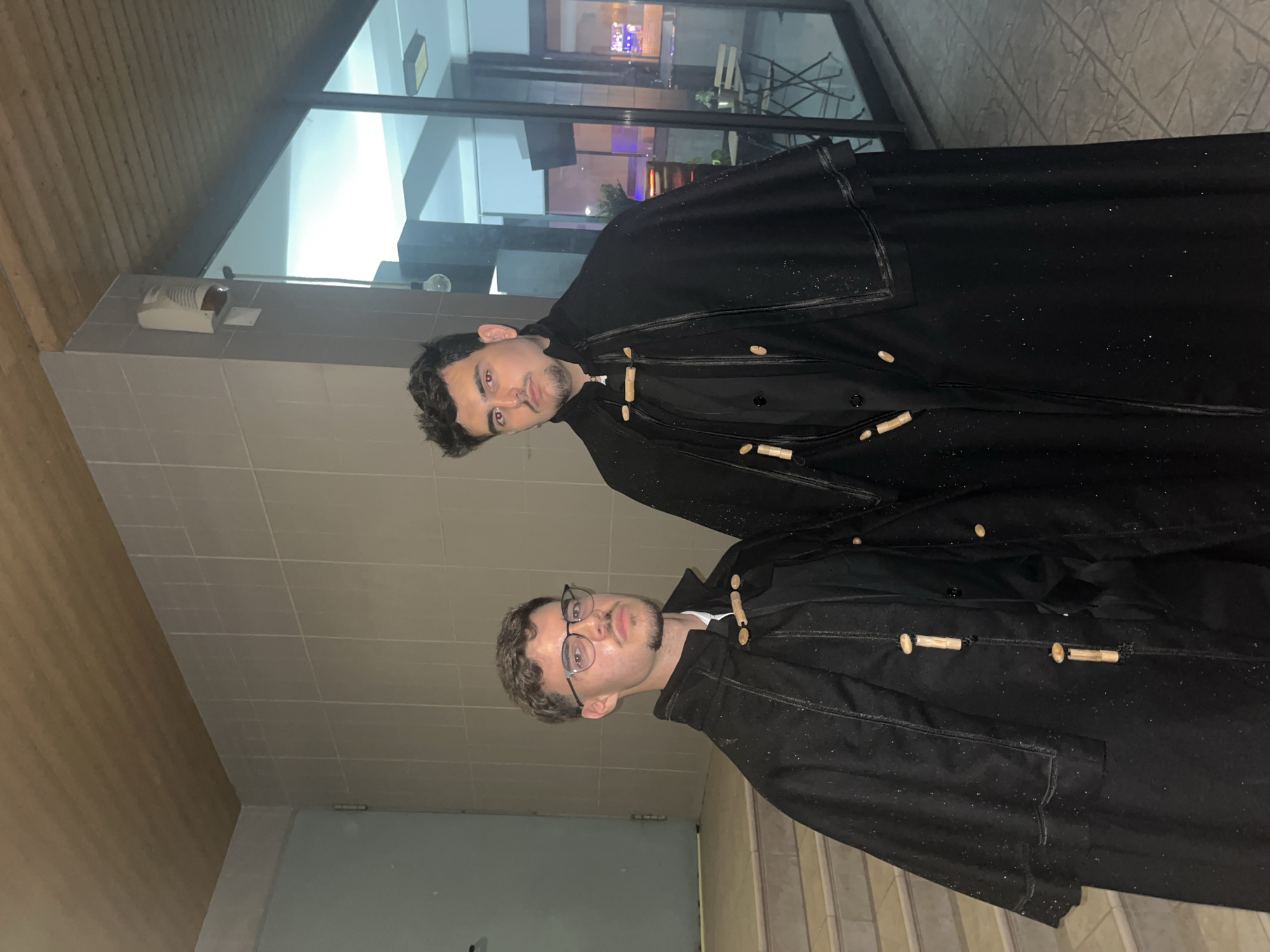 Two young men wearing black cloaks or jackets with wooden toggle fasteners stand and gaze directly at the camera. The image is taken in what appears to be an indoor or covered outdoor passage, possibly at night. The man on the left has dark hair, a short beard, and exhibits red eyes, likely due to camera flash reflection. The man on the right, also with dark hair and a short beard, wears glasses. Both men maintain neutral expressions.
The setting features light grey tiled walls on the left, a wooden-paneled ceiling, and large glass windows or panels behind the subjects. These reflective surfaces show distorted images and reveal an exterior area with artificial lighting, consistent with an evening or nighttime scene in Vila Real, Portugal. A white wall-mounted dispenser is visible in the background on the left. Small white flecks, possibly dust or lint, are scattered across the black fabric of their clothing. No visible text is present in the image.