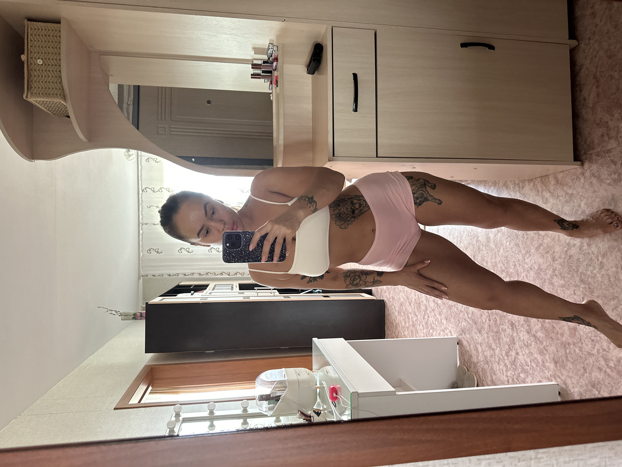 The image shows a young woman taking a mirror selfie in what appears to be a bedroom or dressing area in Yuzhnouralsk, Russia. She is wearing a white sports bra and pink shorts, and has tattoos on her legs and torso. She is holding a smartphone in her right hand, and her left hand is resting on her left thigh.
The room is decorated in a feminine style, with pink floral patterned wallpaper and a pale pink carpet. To the left of the woman is a vanity table with a mirror, makeup, and a pink charging cable. To her right is a light-colored wooden wardrobe with a mirror and several drawers. On the top shelf of the wardrobe, there is a woven basket. The overall impression is one of a personal and well-maintained living space.
