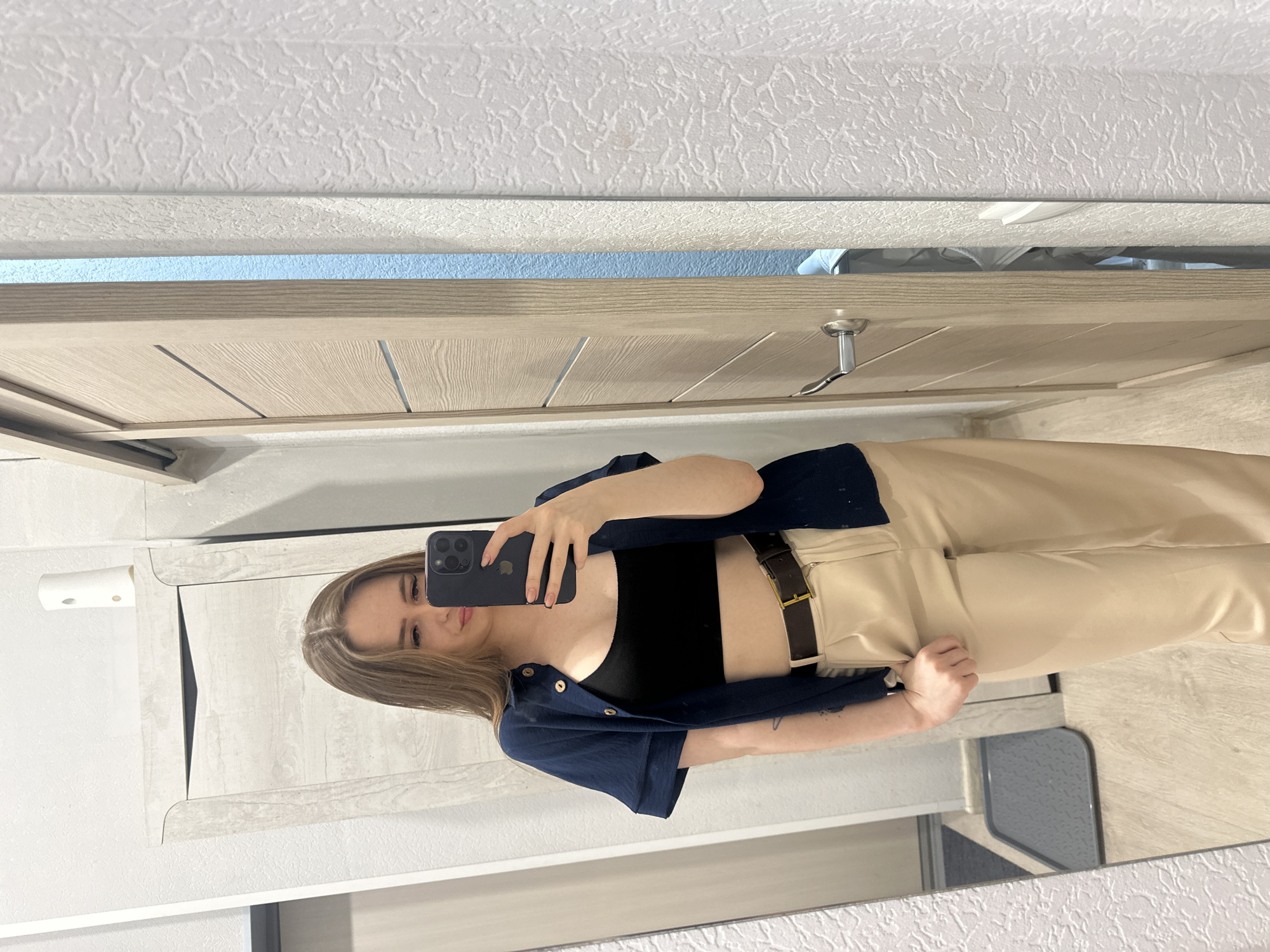 A young woman poses for a selfie in a bathroom mirror. She is wearing a black crop top, beige trousers with a dark brown belt, and a navy button-down shirt worn open. She holds a black smartphone in her right hand to take the photo. The bathroom features a light wooden door with a silver handle, light gray walls, and a mirror with a patterned frame. The location is Unknown, Unknown.