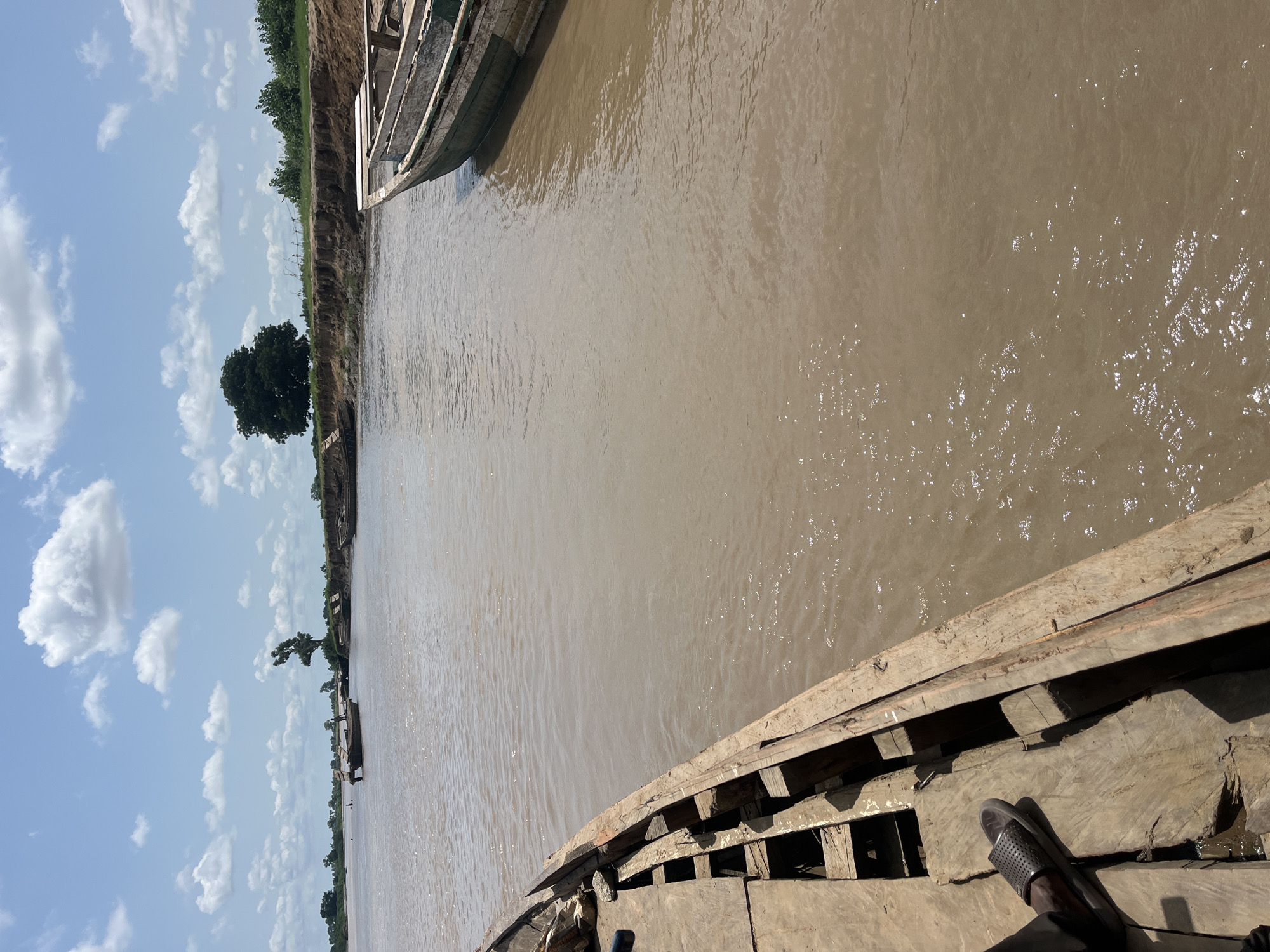 The image depicts a wide, muddy river in Nemnei, Nigeria, under a bright, cloudy sky. In the foreground, the wooden interior of a boat is visible, with a person's left leg wearing a black sandal resting on the wooden planks. The boat appears to be moored or moving slowly.
The river itself is a dominant feature, with its murky brown water reflecting the sunlight. On the far bank, a steep, eroded clay embankment rises, topped with green vegetation and a few scattered trees. The most prominent tree on the far bank is a large, dark-leafed specimen situated on a bend in the river.
In the distance, several other wooden boats are visible, some of them small and flat-bottomed, suggesting local transportation or fishing activities. The overall scene conveys a sense of rural life and the importance of the river as a mode of transit and sustenance in the area. The environment appears natural and undeveloped, with no man-made structures visible other than the boats themselves and possibly some distant, indistinct forms that could be part of the landscape or very distant structures.