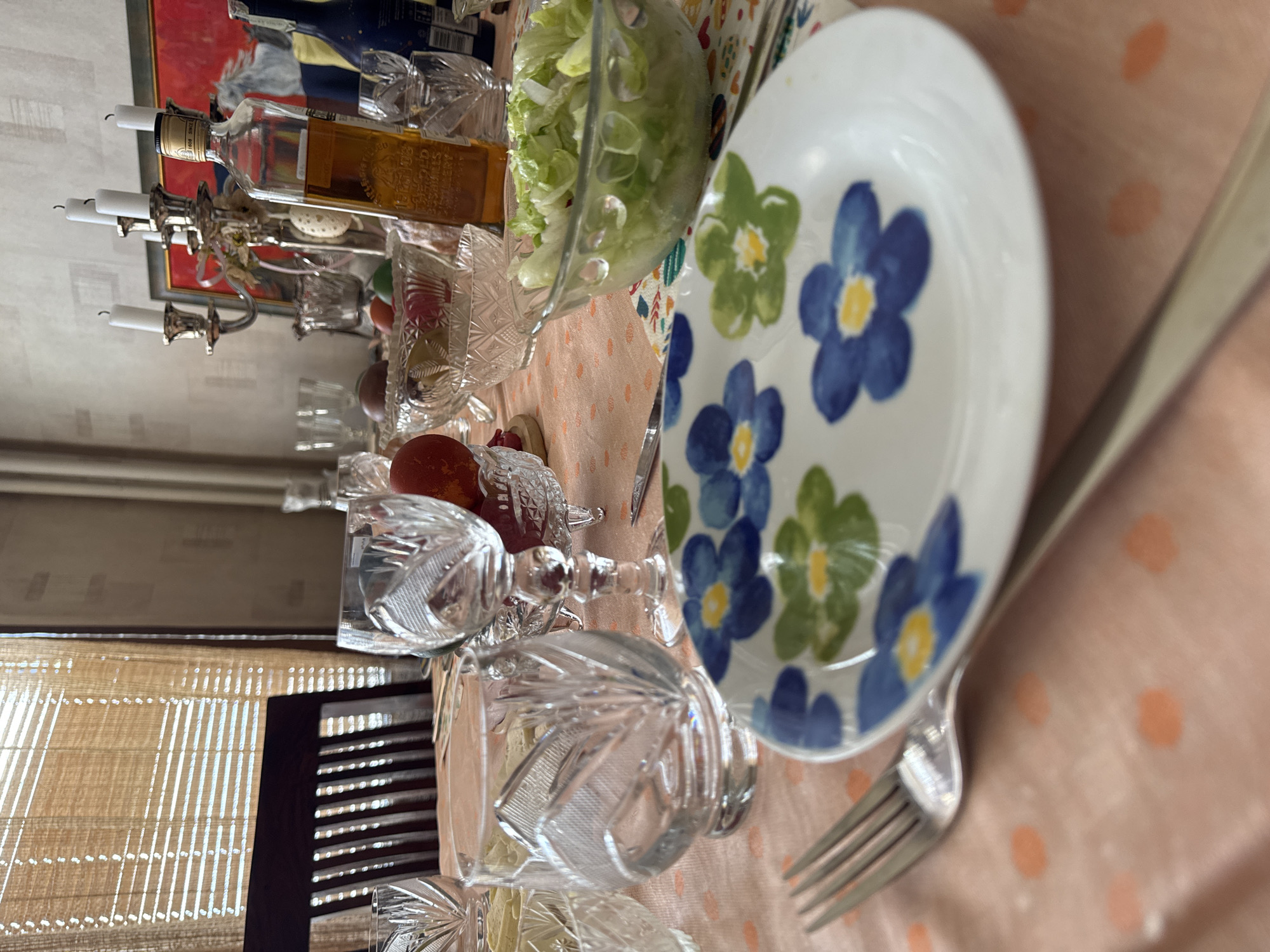 The image shows a dinner table set with a floral plate, a fork, crystal glasses, a bowl of salad, and a bottle of liquor. A candelabra with candles sits on the table. The scene is indoors, near a window with blinds. The location is in Sofia, Bulgaria.