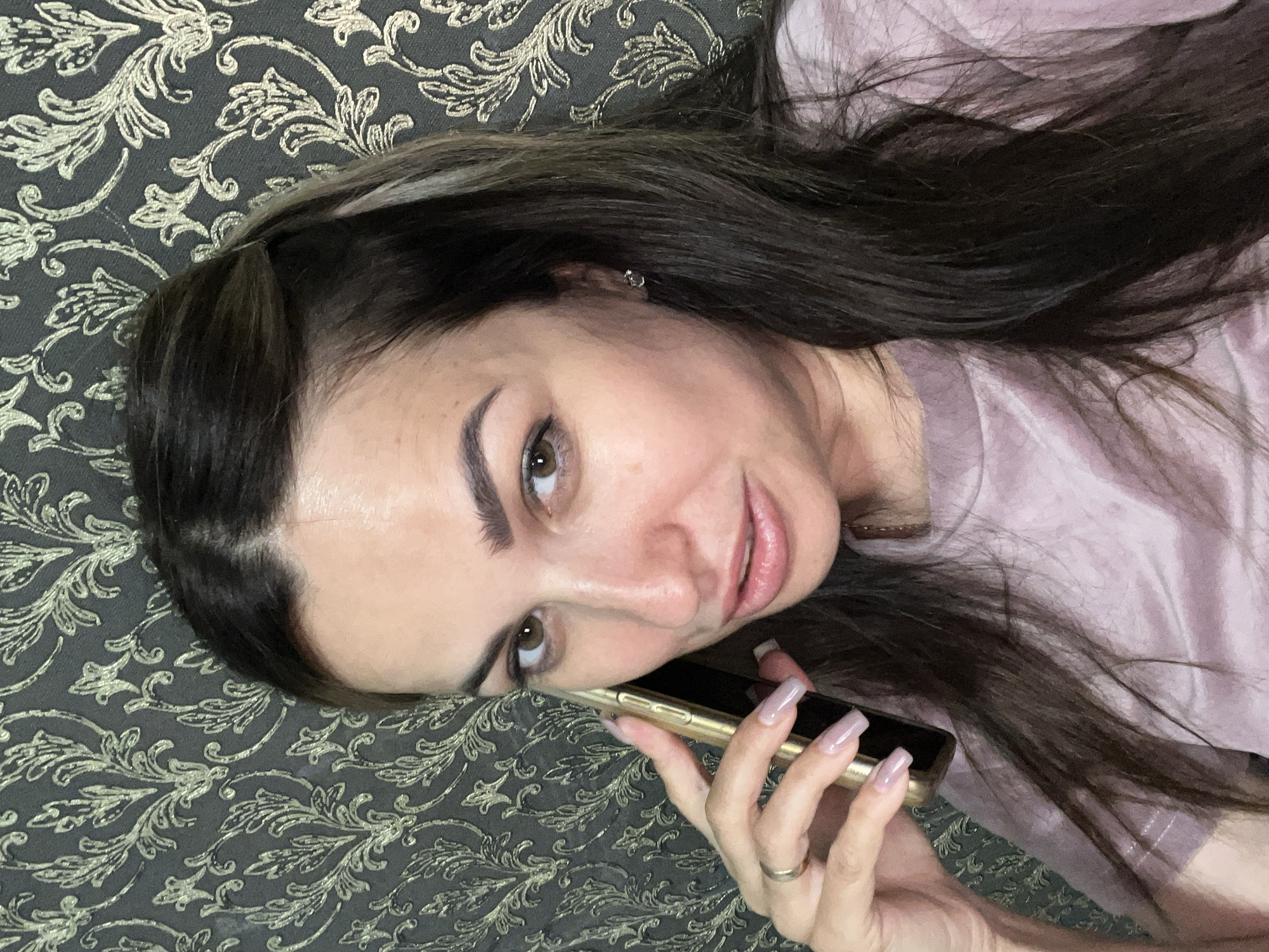 A close-up, waist-up indoor portrait shows a woman with long, dark hair and brown eyes holding a gold smartphone to her left ear with her right hand. She is wearing a light pink t-shirt. Her left hand is visible near her face, and she is wearing a ring on her left ring finger. The background is a dark patterned wallpaper with gold accents. The lighting is soft, and the woman is looking directly at the camera with a slightly parted-lip expression.