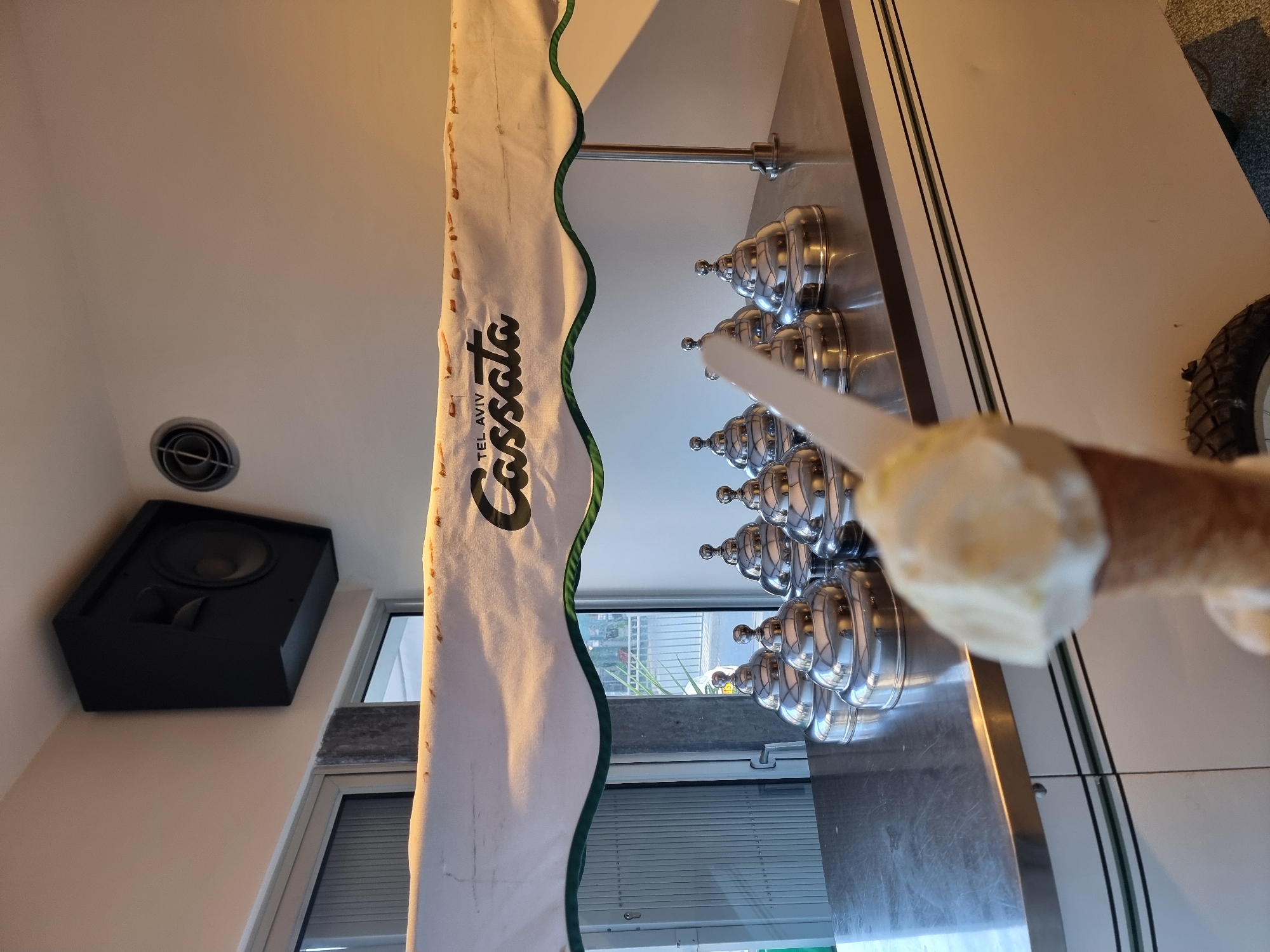 The image depicts a gelato display case with the "Cassata Tel Aviv" logo. The case features rows of metallic gelato containers. In the foreground, a hand holds a gelato cone. The location is identified by the logo as Tel Aviv, Israel. To the left, a black speaker is mounted on the wall near a window.