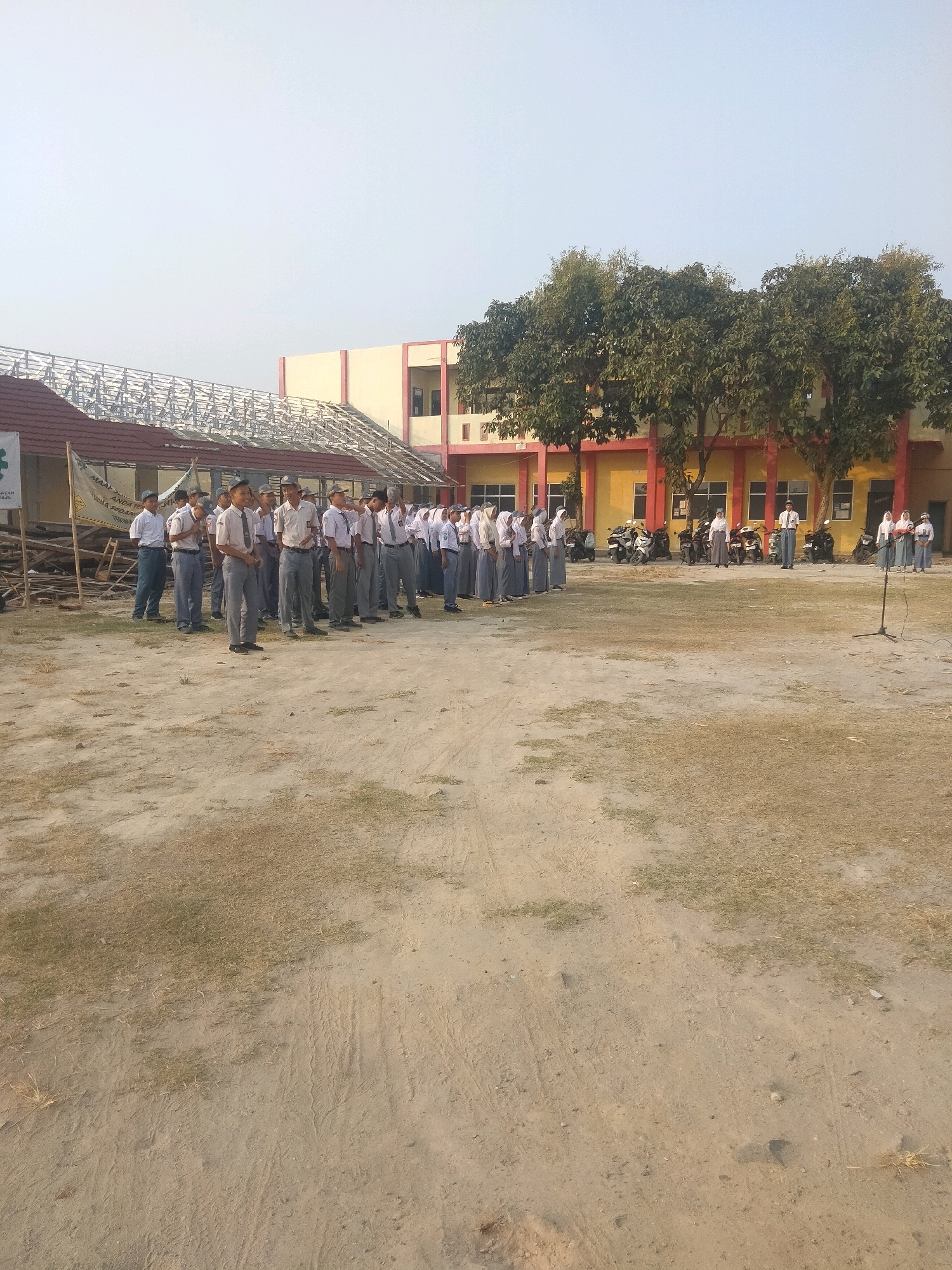 In Suka Sari, Indonesia, a group of students in uniform stand in a formation on a dirt and grass field in front of a two-story building with yellow and pink accents. The building has several windows and doors, and motorcycles are parked in front of it. Trees with full green foliage are situated between the building and the students. In the foreground, a microphone on a stand is positioned in front of the assembled students. The sky is clear and bright, suggesting daytime. Some students are wearing white shirts and grey pants, while others are wearing headscarves and long skirts, indicating a co-educational setting. The scene appears to be an outdoor assembly or event.
