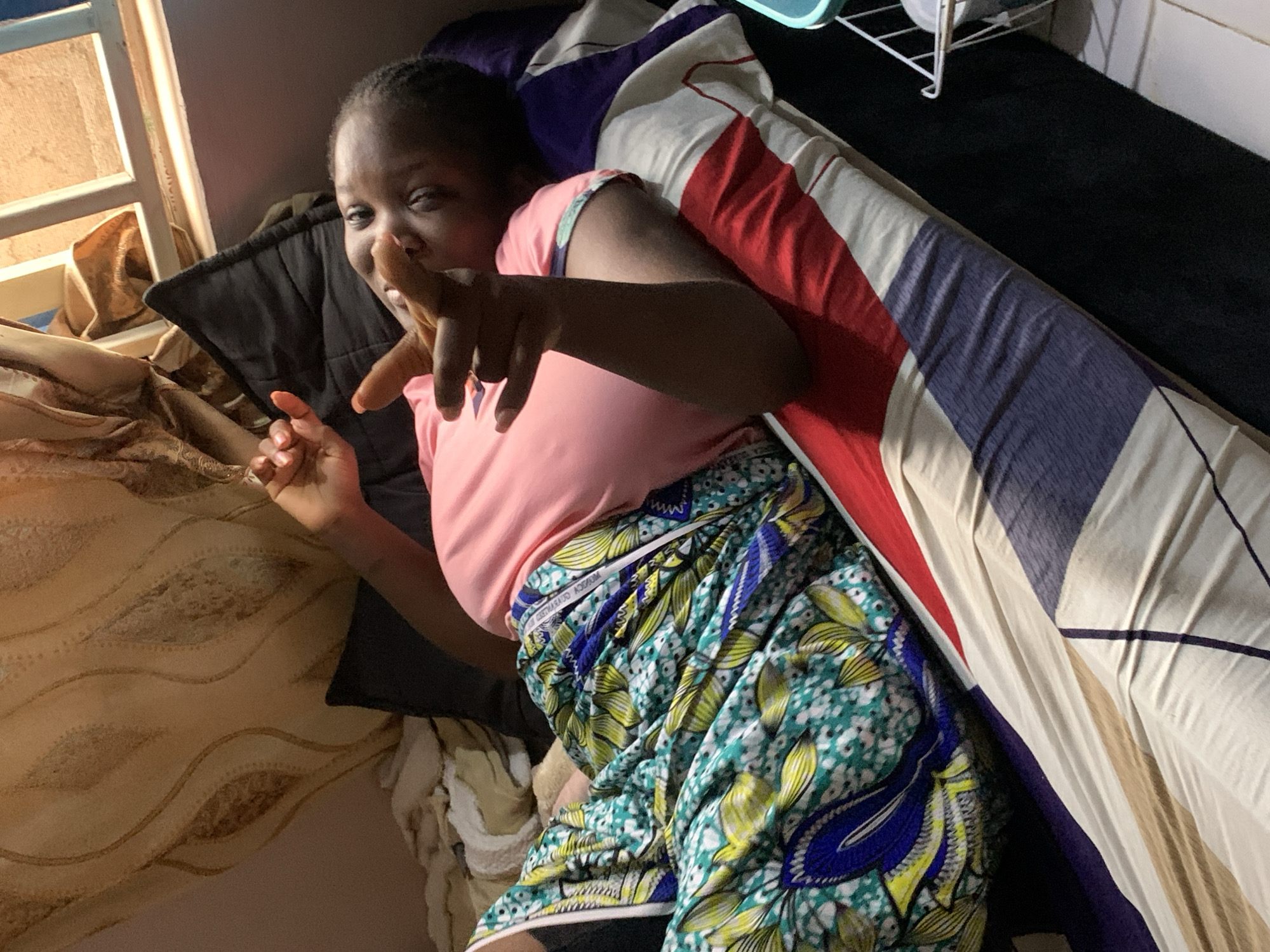 A person is lying on a bed indoors. The person is wearing a pink shirt and patterned shorts with green, yellow, and blue designs. They are gesturing with their fingers. The bed has pillows and blankets with varying patterns. A window is to the left, and part of a shelving unit is visible in the upper right. The image was taken in Rafin Bauna, Nigeria.