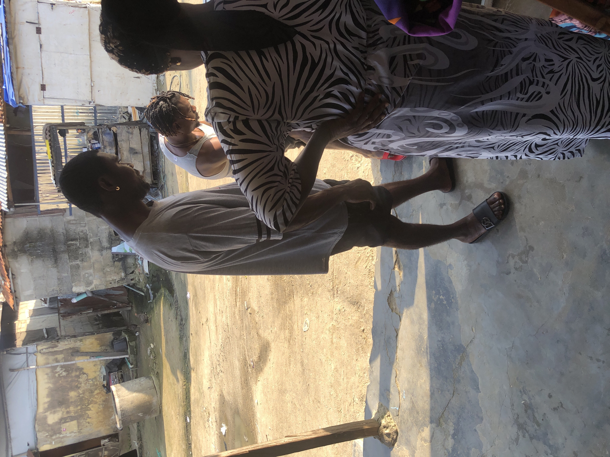 The image depicts an outdoor scene in Ado, Nigeria, under bright sunlight. In the foreground, three individuals are visible. A man on the left, facing away from the camera, wears a grey t-shirt and dark shorts, with a gold hoop earring in his visible ear. He is wearing black sandals. In the center, a person wearing a white tank top is looking downwards. To the right, a person, seen from the back, is wearing a dress with a black and white zebra-like pattern. This individual has their left hand on their hip.
The environment appears to be a somewhat unpaved area, possibly a street or courtyard, with patches of dirt and some puddles of water or mud. In the background, there are low-rise structures with weathered walls, some made of concrete blocks and others appearing to be corrugated metal or makeshift materials. A large concrete cylindrical object, resembling a well or water tank, is present on the left side of the background. There are also chickens visible in the background, one of which is pecking at the ground. The overall impression is of a humble, possibly developing urban setting.