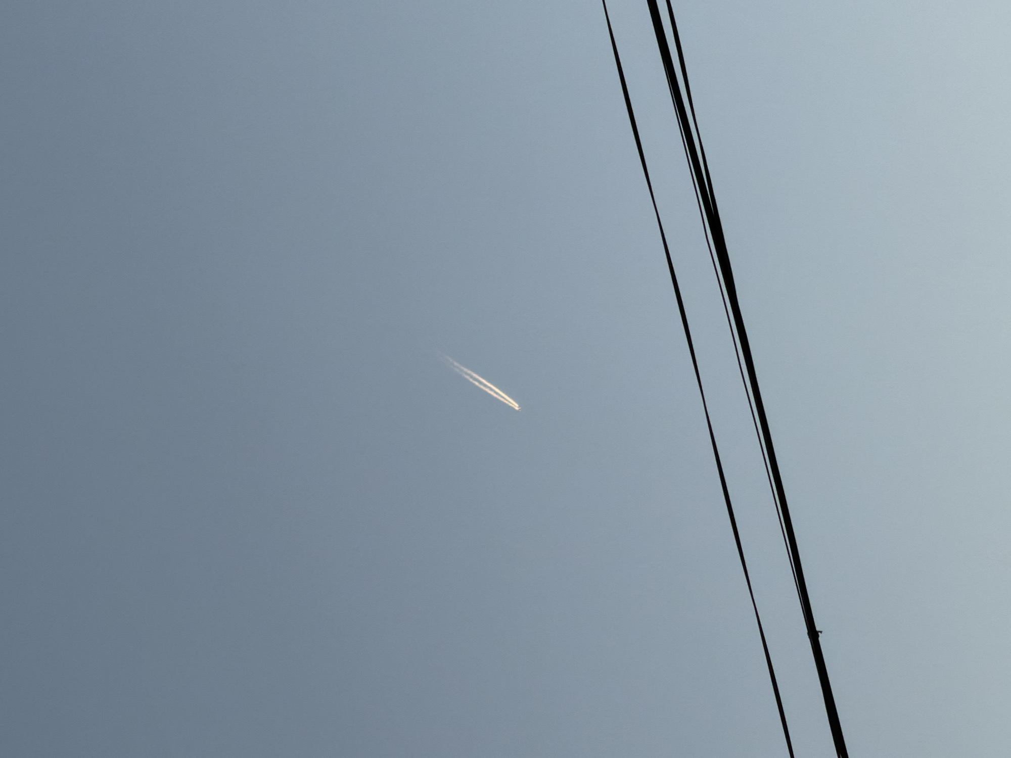 The image shows a blue sky with a contrail from an airplane. Electrical wires are visible in the right side of the frame. The location is unknown, and no people, objects, actions, or interactions are discernible beyond the sky, contrail, and wires. The environment is a clear sky.