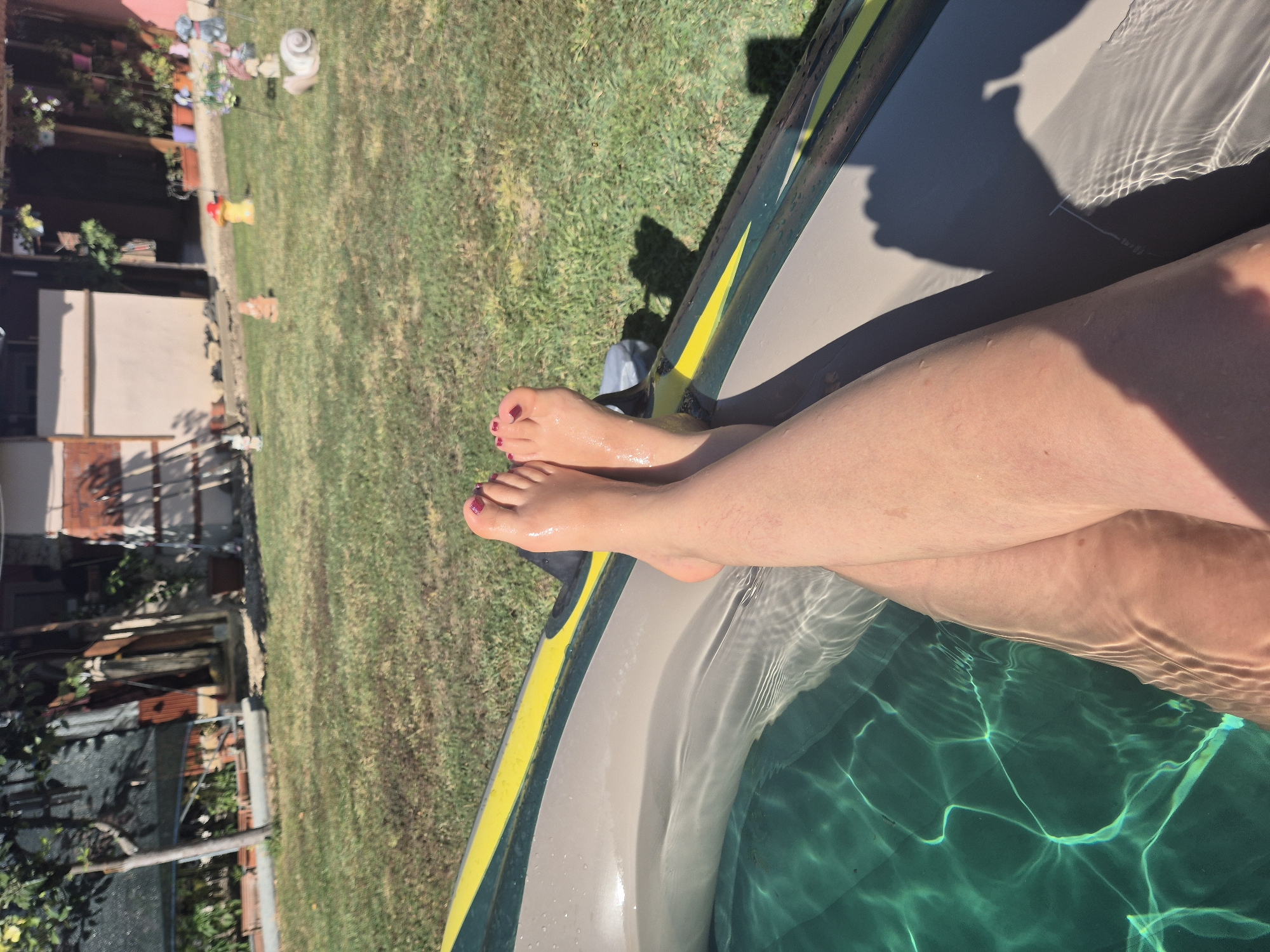 A close-up, aerial shot shows a person's legs and feet extending from the right side of the frame into a partially submerged inflatable pool, which is filled with clear, green water. The pool has a grey and yellow rim. The person's feet are wet and have painted toenails. The background consists of a grassy yard with scattered garden decorations and a building. The sun casts shadows from the pool and unseen objects onto the grass.