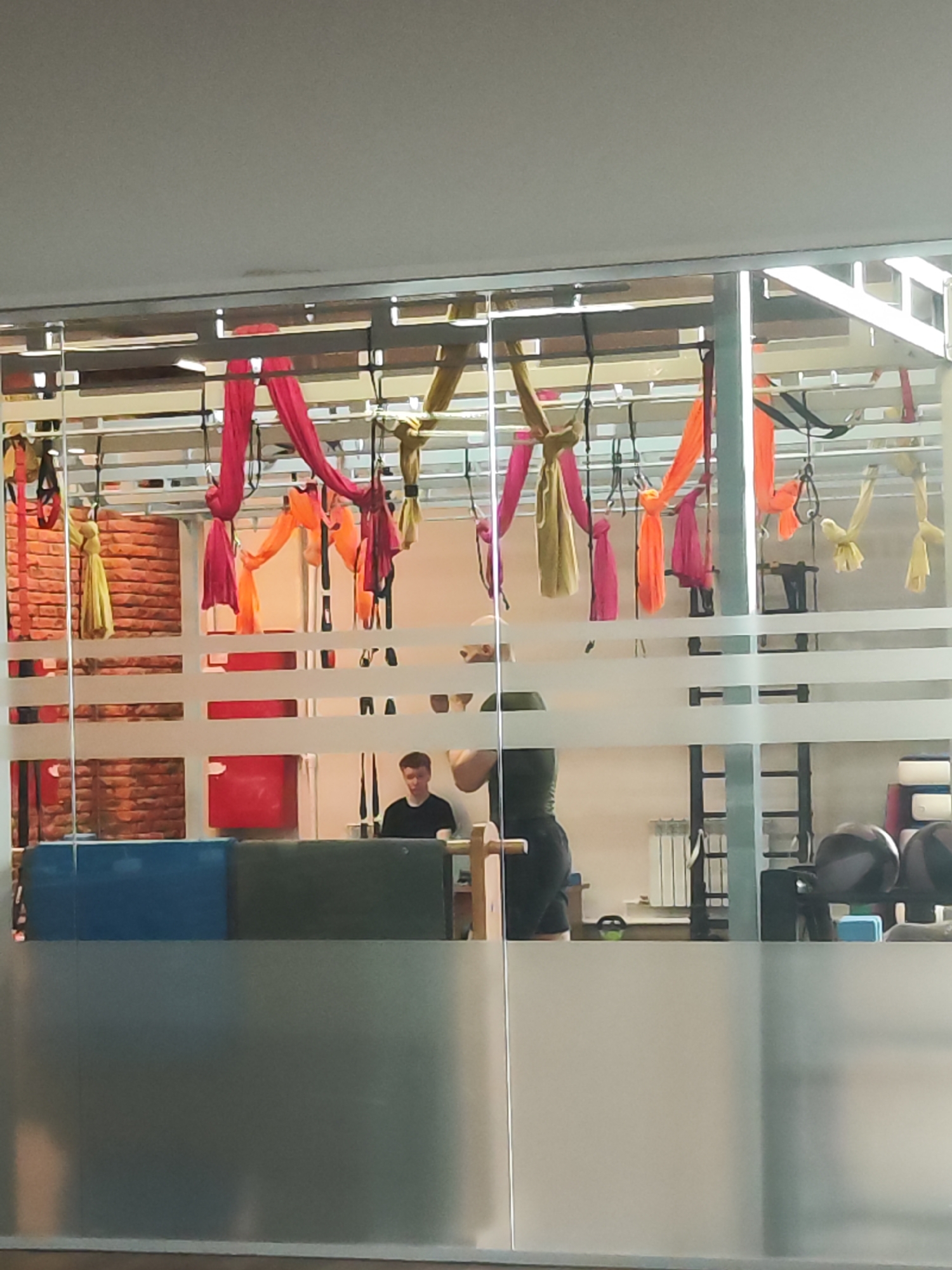 The image, taken from outside a building in Chaikovsky, Russia, shows the interior of a fitness studio. The view is partially obscured by frosted glass. Inside, there is a rig supporting aerial silks in various colors: yellow, orange, and pink. Two people are visible: one seated at a desk, and another standing. Exercise equipment, including free weights and a ladder, is in the background. The studio has a brick wall.