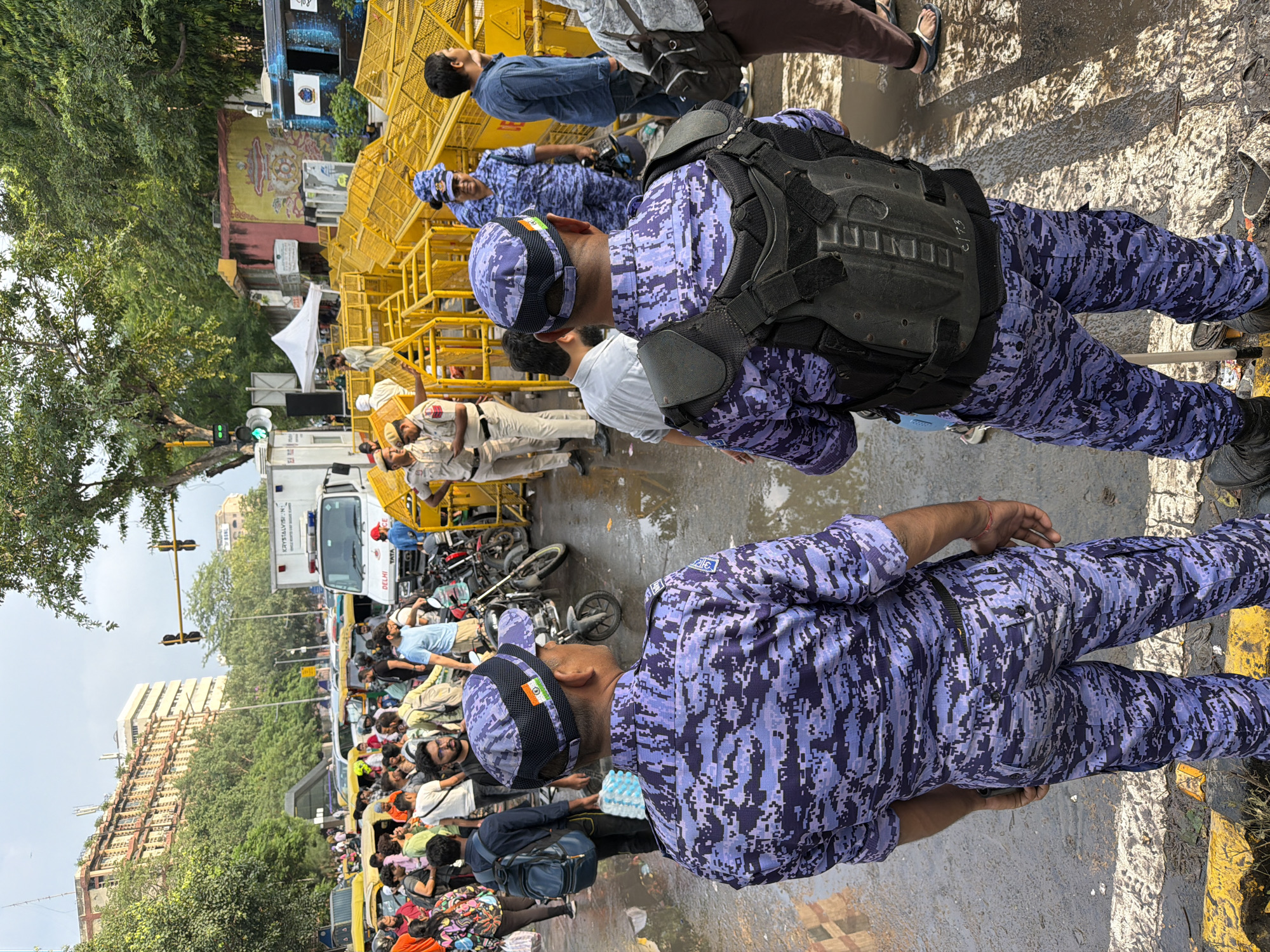 The image depicts a street scene in New Delhi, India, likely during daytime given the lighting conditions and overcast sky. The foreground features two individuals in distinct purple camouflage uniforms, identified as security personnel. The individual on the left faces away from the camera, wearing a cap with an Indian flag patch. The person on the right also faces away, with a tactical vest over their uniform. Both are standing on a wet, asphalt road, marked with yellow lines.
The middle ground shows a crowd of people, some in civilian attire, others in similar uniforms. There are yellow metal barricades forming a barrier, and behind them, a white truck with "TATA" visible on its side, possibly a police or emergency vehicle. Several motorcycles are parked in the area. In the background, a multi-story building with arched windows and some trees are visible, suggesting an urban environment. The overall scene appears to be one of crowd control or a public event with a significant security presence.