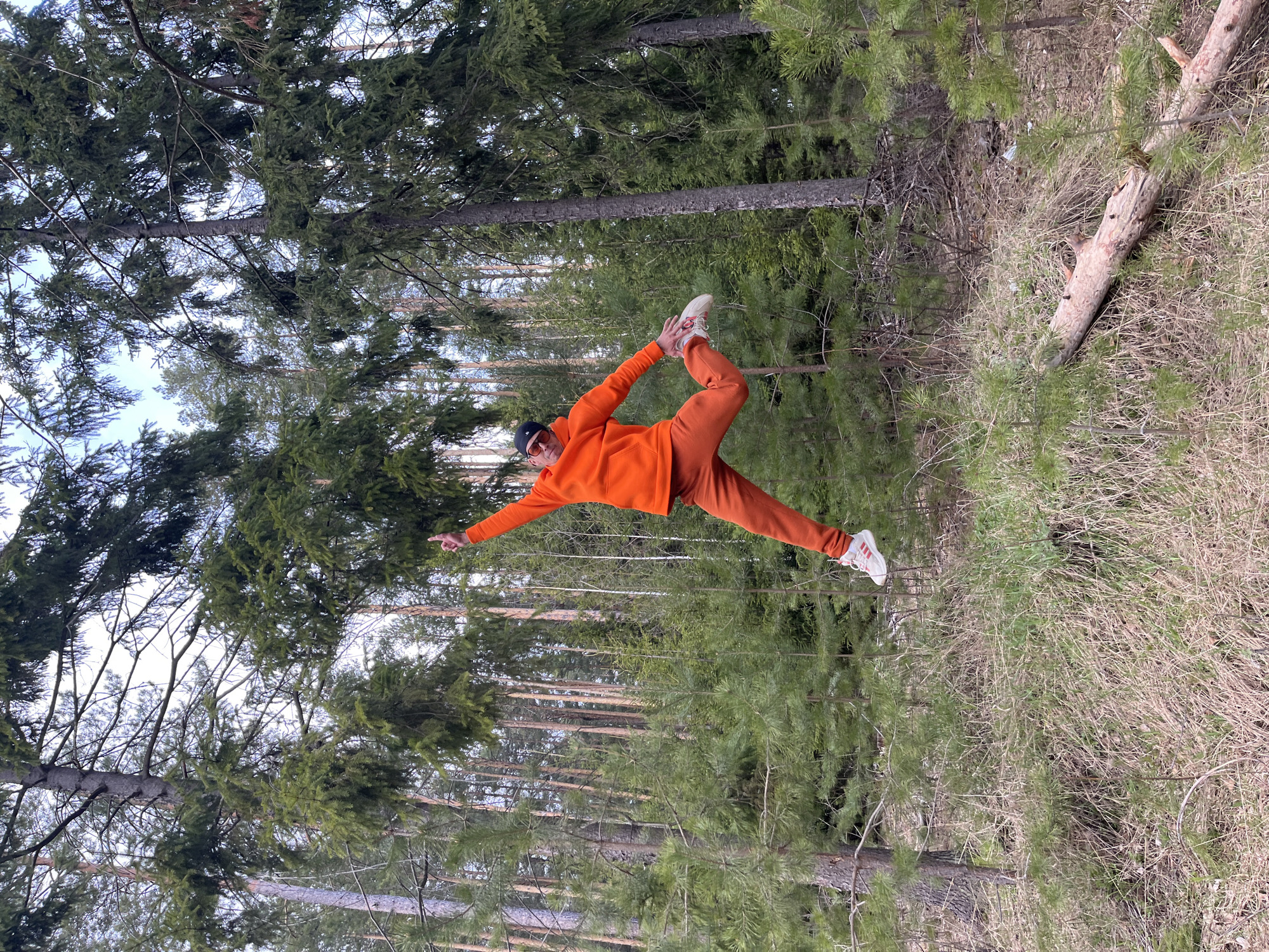 An individual is captured in a full-body shot, performing a dynamic balancing pose in a forest setting in Chaikovsky, Russia. The person, appearing male, is dressed in a vibrant, monochromatic orange outfit consisting of a hoodie and sweatpants, paired with white sneakers with red accents and a dark baseball cap. They are seen balancing on one leg, with the other leg bent backward and its foot held by the corresponding hand, while the opposite arm is extended forward and pointing. This pose resembles a yoga or dance stance, possibly a variation of the Dancer's Pose.
The setting is a coniferous forest, likely composed of pine or spruce trees. Tall, slender tree trunks rise from the ground, topped with dense green foliage that partially obscures a bright, clear sky. The forest floor is covered with dry, light-brown grass and fallen pine needles, with some patches of greener undergrowth. A large, weathered log lies horizontally on the right side of the frame, blending into the natural environment. The lighting suggests it is daytime, possibly late morning or early afternoon, with clear weather. The bright orange clothing creates a strong contrast with the natural greens and browns of the forest, making the subject stand out. There is no visible text in the image.