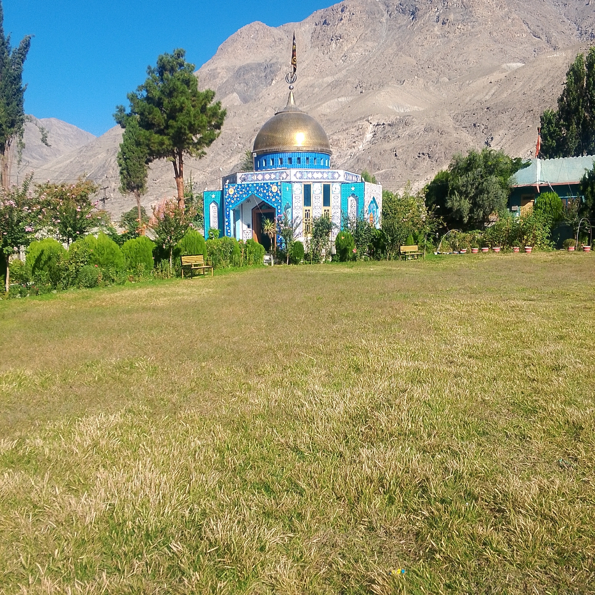 This image depicts a mosque with a golden dome, situated in a mountainous landscape, possibly in the city of Gilgit, Pakistan. The mosque features intricate blue and white tile work. In front of the mosque, there is a well-maintained green lawn with some trees and bushes. Two wooden benches are visible on the lawn. The background is dominated by a large, barren mountain range under a clear blue sky. The overall scene conveys a sense of peace and natural beauty, characteristic of a mountainous region.