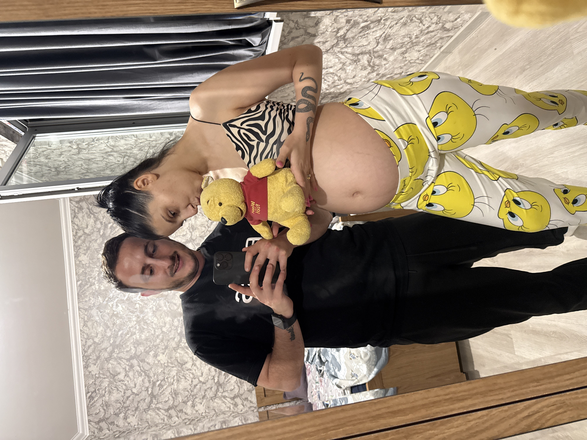 In a mirrored reflection, a man and a pregnant woman are seen in what appears to be a residential interior, possibly in Tbilisi, Georgia. The man, on the left, wears a black t-shirt and black pants, holding a smartphone to take the selfie. He has a short beard and is smiling. The woman, on the right, is pregnant and wears a zebra-print camisole and white pants adorned with cartoon Tweety Bird characters. She is holding a Winnie the Pooh stuffed toy, kissing its head. The background shows a bedroom with a bed and a window covered by gray curtains. The wall is wallpapered with a textured pattern.