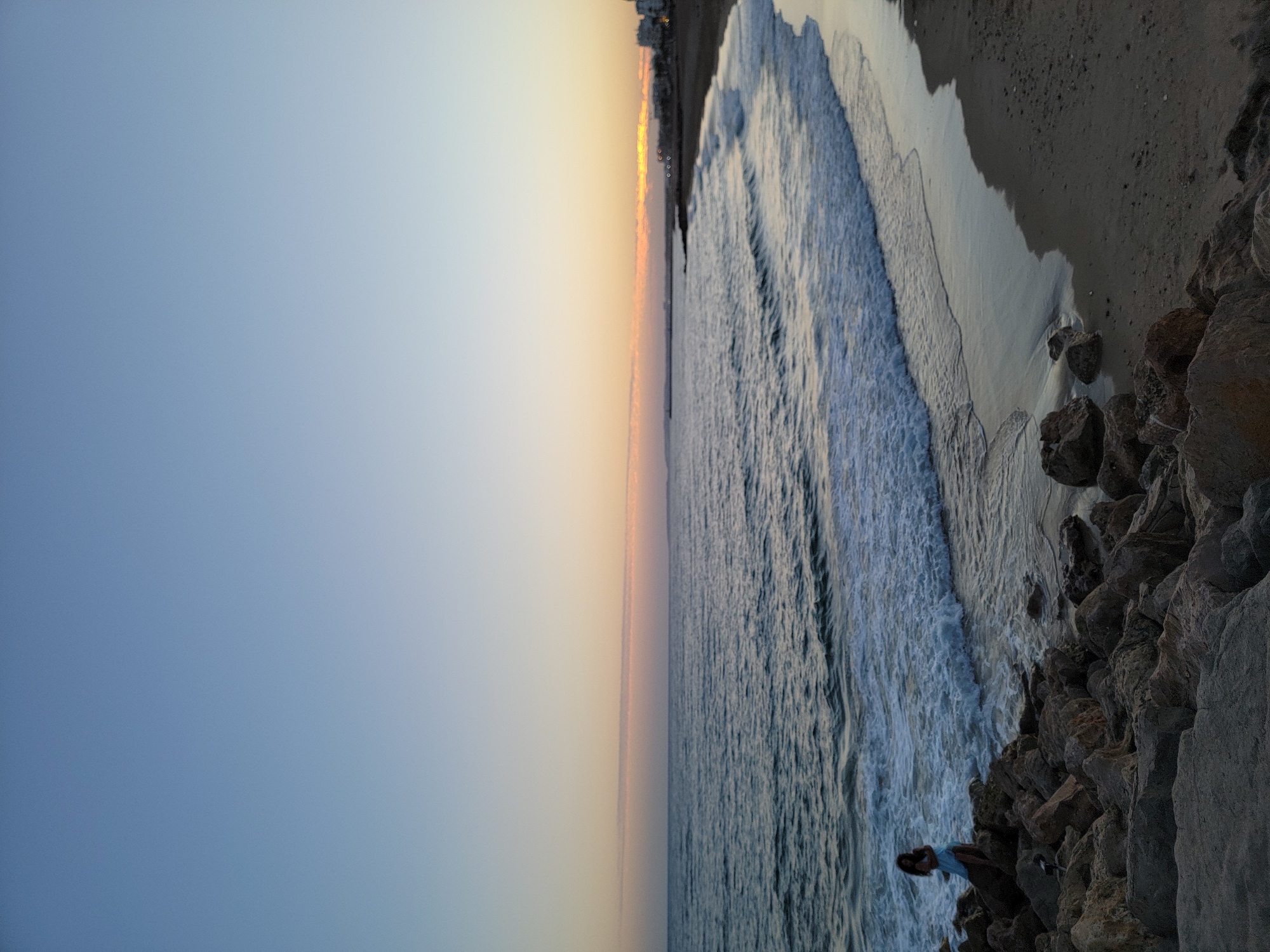An evening scene at Praia de Quarteira, Portugal, showcases the tranquil sea and sky at sunset. The ocean, a deep blue, is dotted with gentle waves that break into white foam as they reach the shore. The beach, a strip of sand, is bordered by a rocky coastline on the left.
On the rocky section, a lone figure, a person wearing a light blue dress, stands facing the sea, their back to the viewer. They appear to be observing the horizon. The sky above is a gradient of soft blues and oranges, transitioning from a clear, pale blue at the top to a warmer hue near the horizon, indicating the sun is setting or has just set. In the distance, along the coast, a line of buildings with lights can be seen. The overall atmosphere is peaceful and serene, capturing the beauty of a coastal evening.