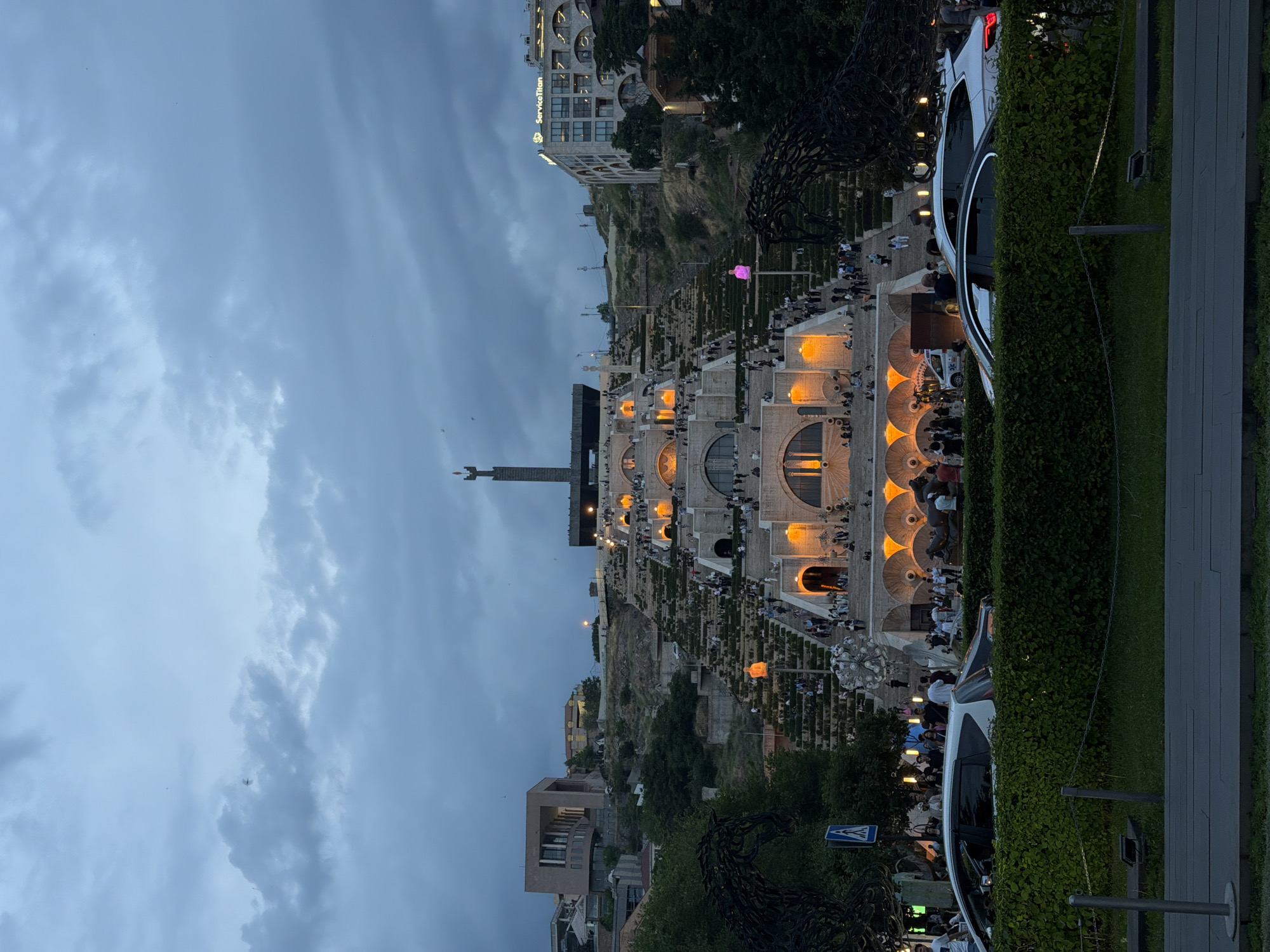 This image depicts an evening scene at a large, illuminated cascading architectural complex under an overcast sky. The prominent structure, characteristic of the Cascade in Yerevan, Armenia, features multiple levels of steps, terraces, and arched openings, all illuminated with warm orange lights. A tall, dark monument stands at the very top of the structure, visible against the twilight sky.
Numerous people are scattered across the steps and terraces, walking, gathering, and enjoying the evening atmosphere. Some appear to be dining or socializing in the illuminated arched sections of the Cascade.
To the right, a road or path is visible with several vehicles, including a white car prominent in the foreground and a partially visible ambulance further back. A dense green hedge separates the pedestrian area from the road. On the lower left, a large, dark, metallic sculpture, resembling a horse, is visible amidst greenery. Higher up on one of the terraces, a distinctive glowing pink/purple human-like sculpture adds a modern artistic touch. Another spherical light sculpture is also present.
Buildings are situated on the hillside surrounding the Cascade. One building on the upper left features the illuminated text "ServiceTitan." A street sign with a pedestrian symbol is also visible on the right. The sky is cloudy with visible aircraft in the distance. The overall impression is of a lively and well-frequented public space during the early evening hours.