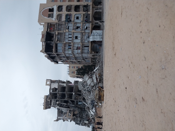 The image shows extensive destruction of buildings, possibly due to conflict or natural disaster. A partially collapsed, multi-story building is visible, with rubble and debris piled up at its base. Nearby, another damaged building stands, displaying signs of significant structural damage, with missing walls and exposed interiors. The ground in the foreground is barren and sandy. Several figures are visible in the bottom corner. The sky is overcast, suggesting a somber atmosphere.