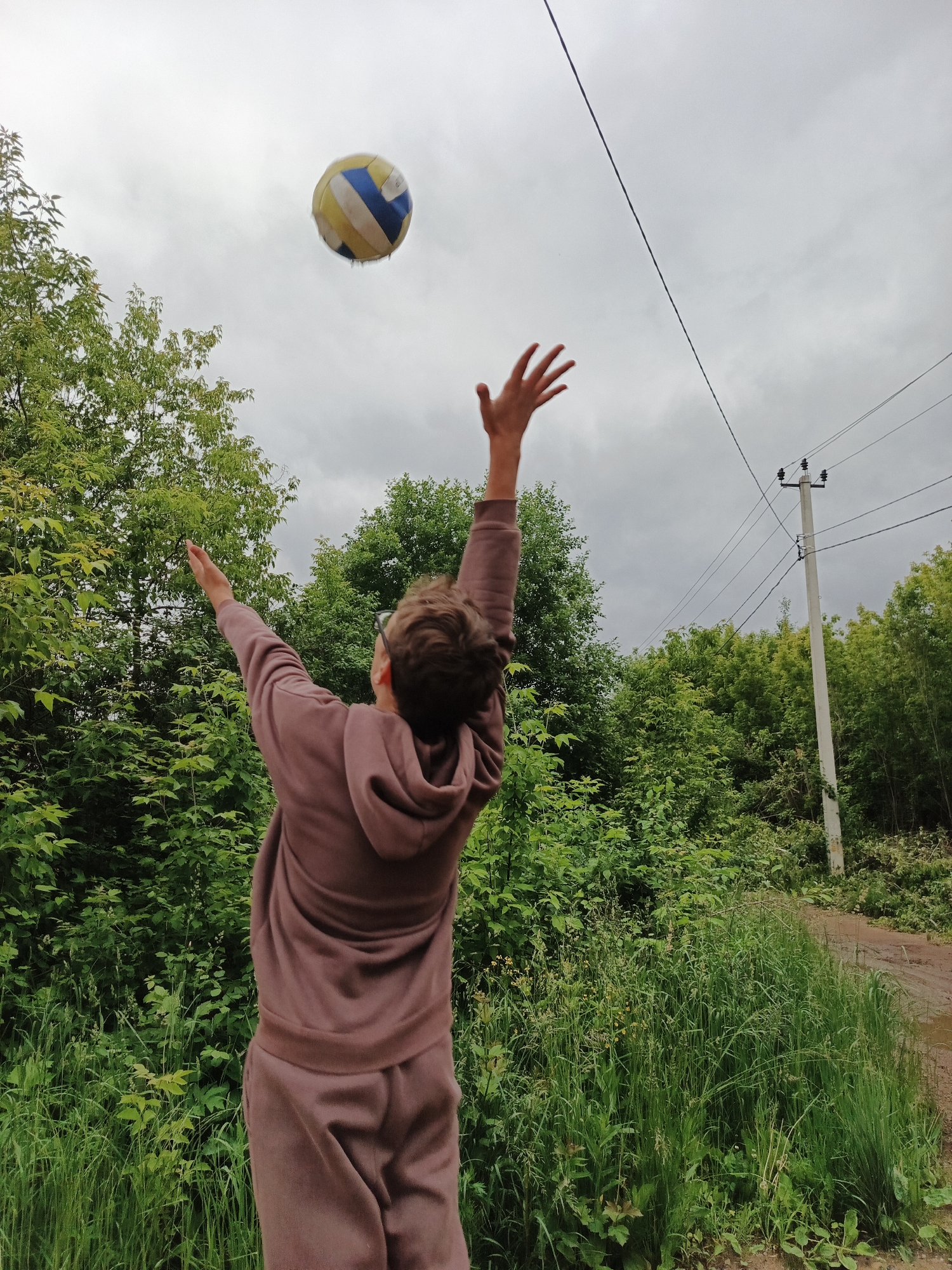 In the city of Unknown, Unknown, a low-angle, eye-level outdoor shot depicts a person in mid-action, attempting to hit a volleyball. The individual, seen from the back, wears a monochromatic two-piece tracksuit in a muted dusty rose or light brown color, consisting of a hooded top and matching pants. Their arms are extended upwards and slightly forward, with palms open, positioned towards a yellow, blue, and white volleyball captured in mid-air slightly above and to the left of their head. The person appears to be wearing glasses, visible behind their right ear.
The environment is characterized by dense green foliage, primarily trees and tall grass, forming a lush backdrop. A dirt path or road is visible on the right side of the frame, partially obscured by the foreground vegetation. The sky is overcast and uniformly light gray, occupying a significant portion of the upper half of the image. A utility pole with multiple black electrical wires extends vertically on the right side of the image, with wires stretching horizontally across the upper third of the frame. The overall impression is an informal outdoor setting, possibly a rural or semi-rural area, during daytime under cloudy conditions.
