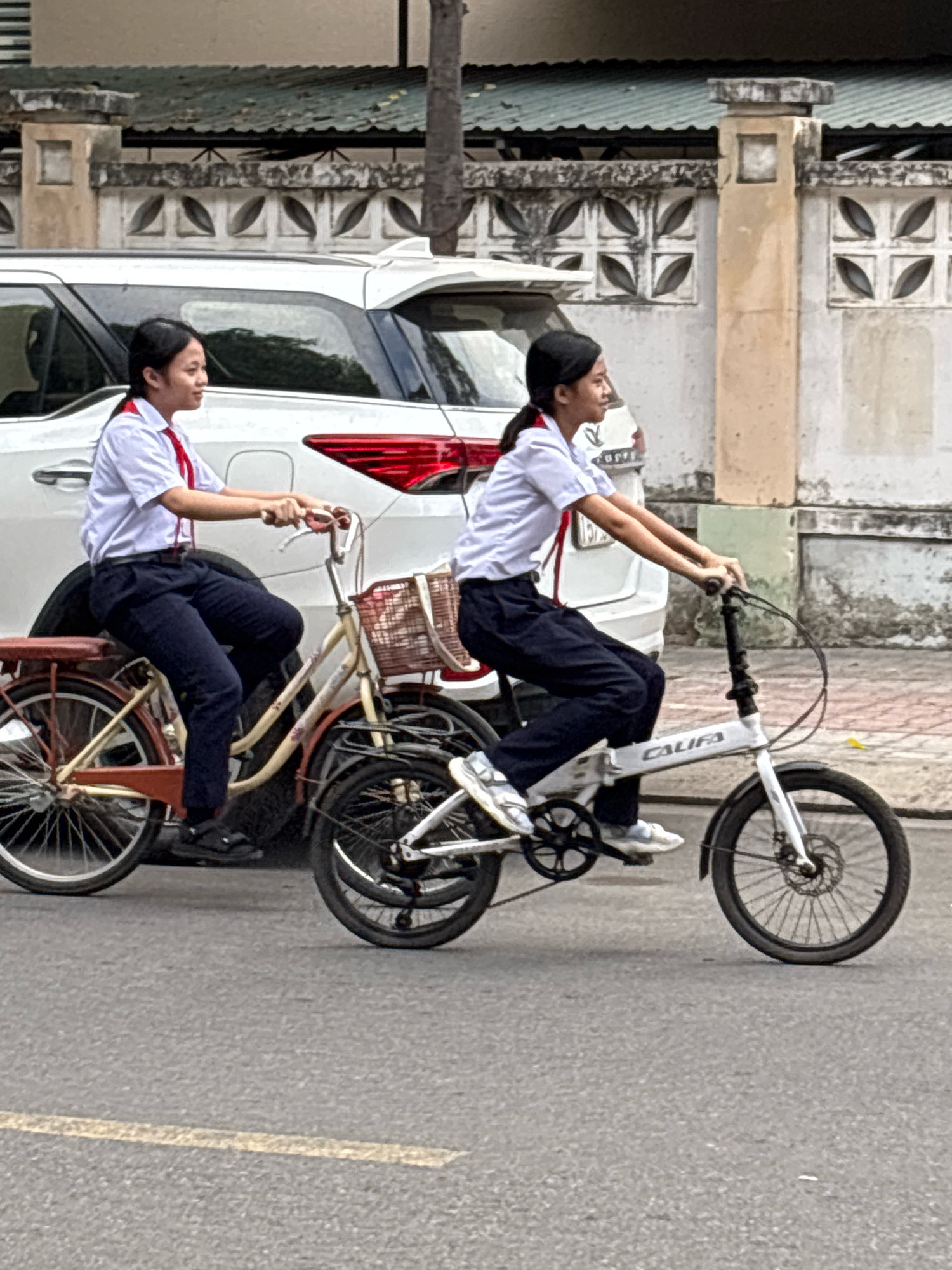 Two young girls in school uniforms, white shirts with red ties and dark pants, are riding bicycles on a paved street. The girl in front is on a white folding bicycle, and the girl behind her is on a larger, older-style bicycle with a basket. Both are looking ahead, and the girl in the front appears to be slightly ahead and to the right of the girl in the back. They are riding next to a white SUV. The background shows a low wall with decorative patterns and a building with a green roof. The scene is consistent with a street in Nha Trang, Vietnam, where students often cycle to and from school.
