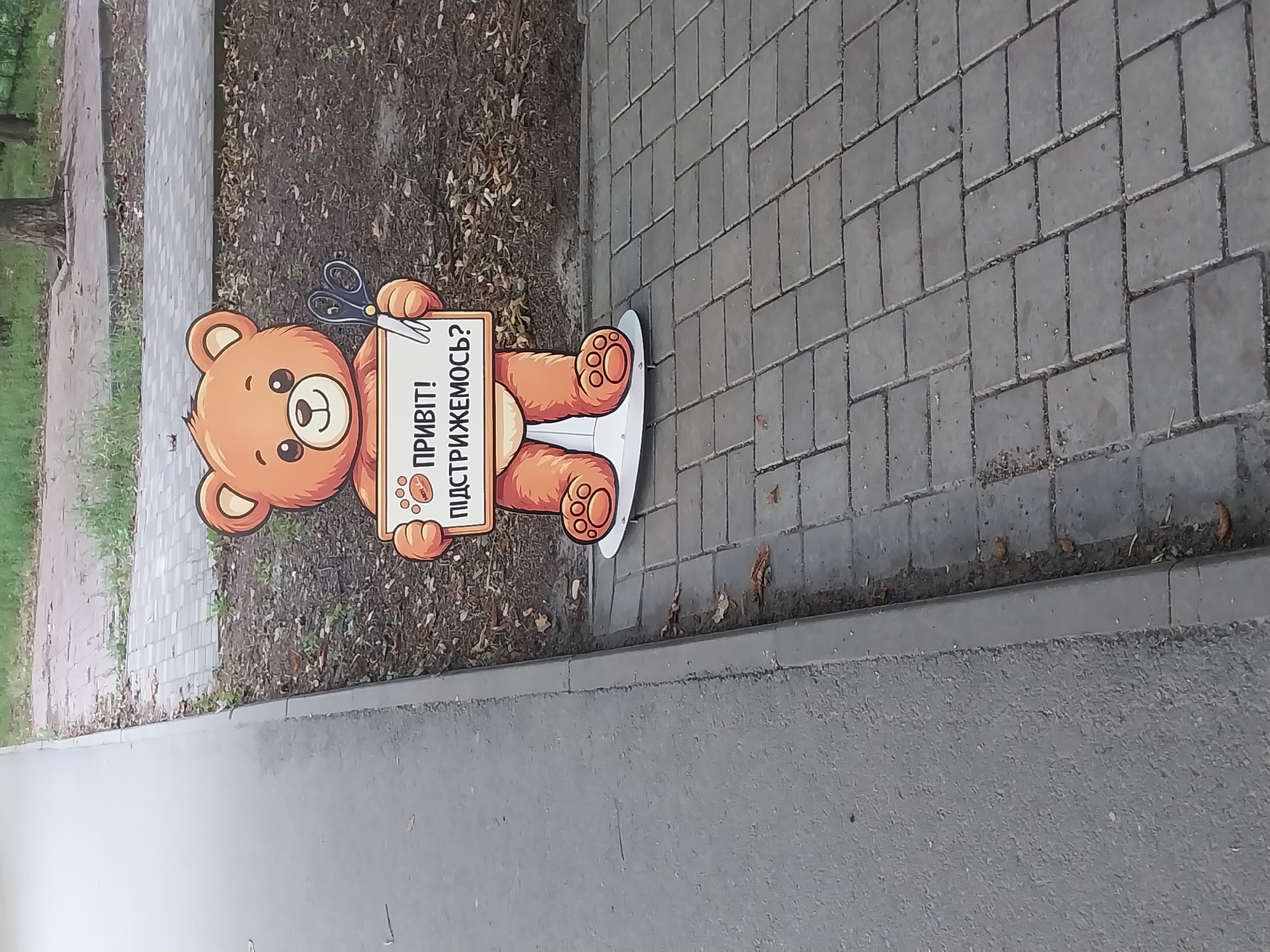 A cardboard cutout of a cartoon teddy bear stands on a paved area. The bear is orange with a white muzzle and holds a white sign with blue text that says "ПРИВІТ! ПІДСТРИЖЕМОСЬ?". It also holds a pair of blue scissors in its right paw. The cutout is positioned on a sidewalk made of grey interlocking pavers, bordering a grey asphalt path. The background consists of dirt with scattered dry leaves and some sparse green vegetation. The scene appears to be outdoors, possibly in a park or along a street in Dnipro, Ukraine.