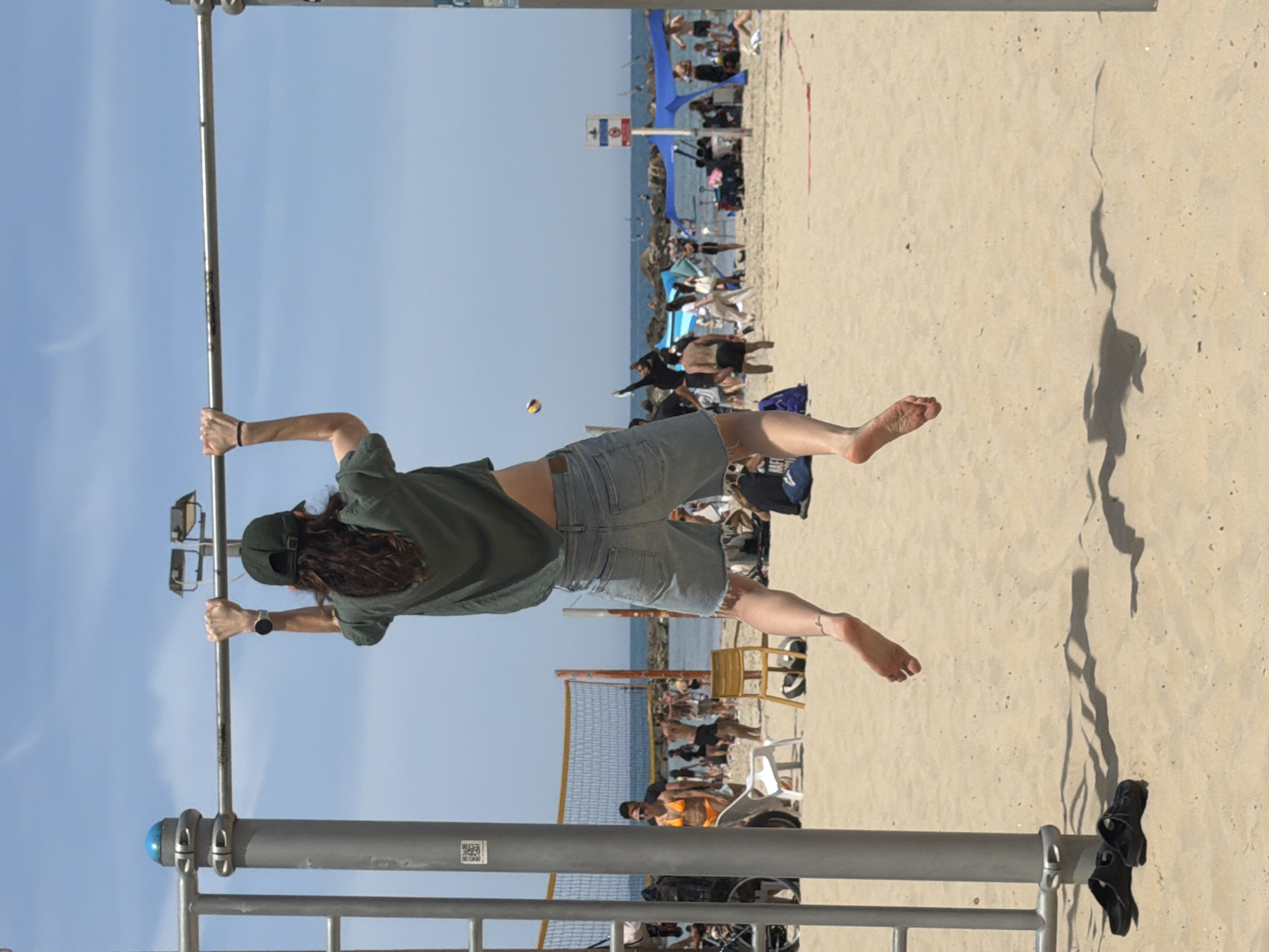 A person hangs from a metal bar structure, seemingly at a beach in Tel Aviv, Israel. The individual wears denim shorts, a green shirt, and a baseball cap. A shadow is cast on the sand near a pair of sandals on a lower metal rail. The beach in the background is populated with people, umbrellas, and a volleyball net. The sky is clear blue.