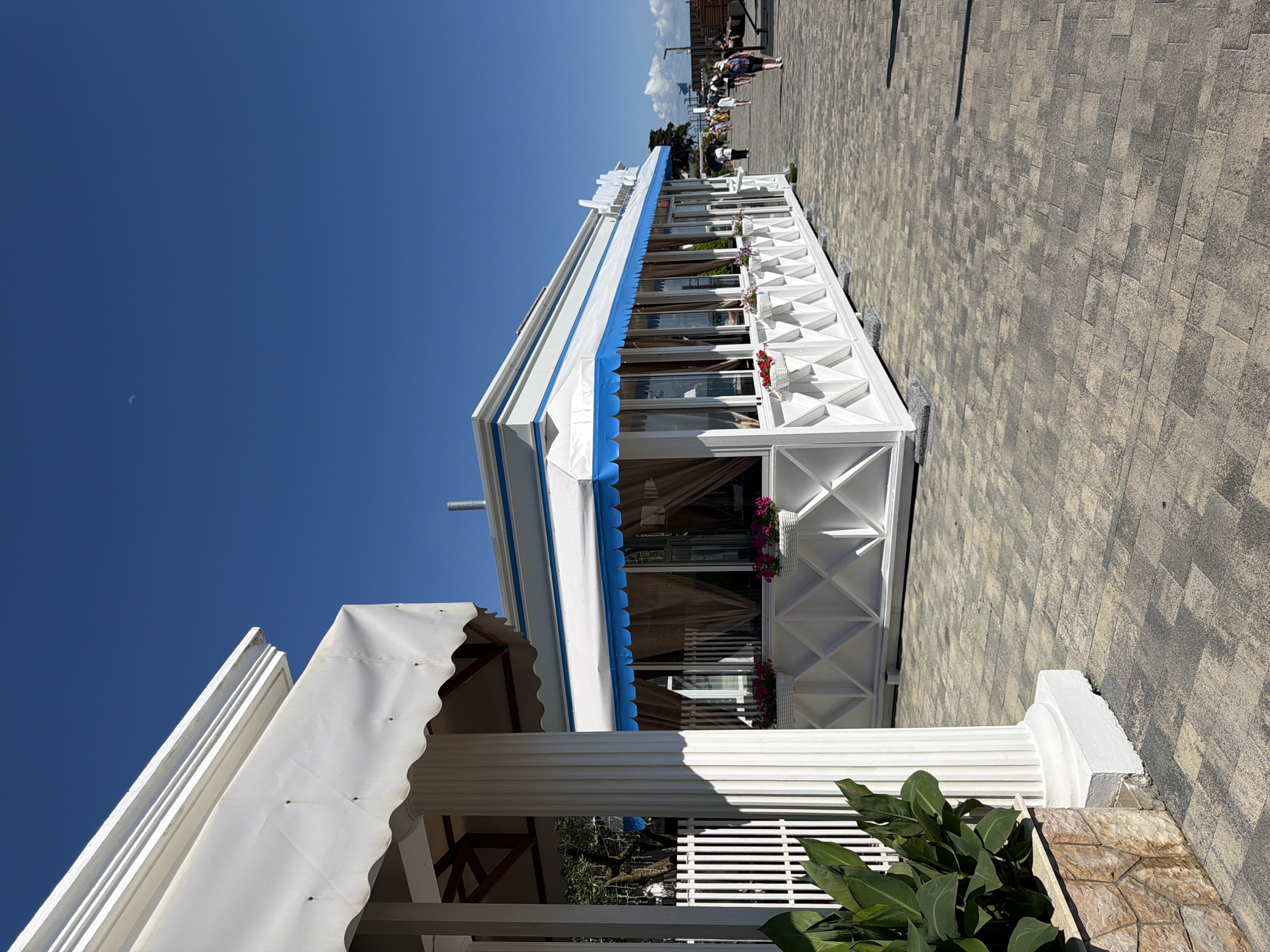 This image captures a sunlit day in Sochi, Russia, likely along the coastline or a promenade. In the foreground, a white building with a blue awning stands prominently. This building appears to be a cafe or restaurant, with large windows and decorative white lattice work around its base, adorned with white flower baskets. To the left, a portion of another white structure with a fluted column and a white awning is visible, suggesting a connected or adjacent establishment.
The ground is paved with light-colored interlocking stones, forming a patterned walkway. Beyond the white building, a clear blue sky stretches overhead, with a faint crescent moon visible. In the background, a few people are walking along the promenade. The overall scene is bright and airy, indicative of a pleasant, warm day. The architecture suggests a resort or leisure area.
