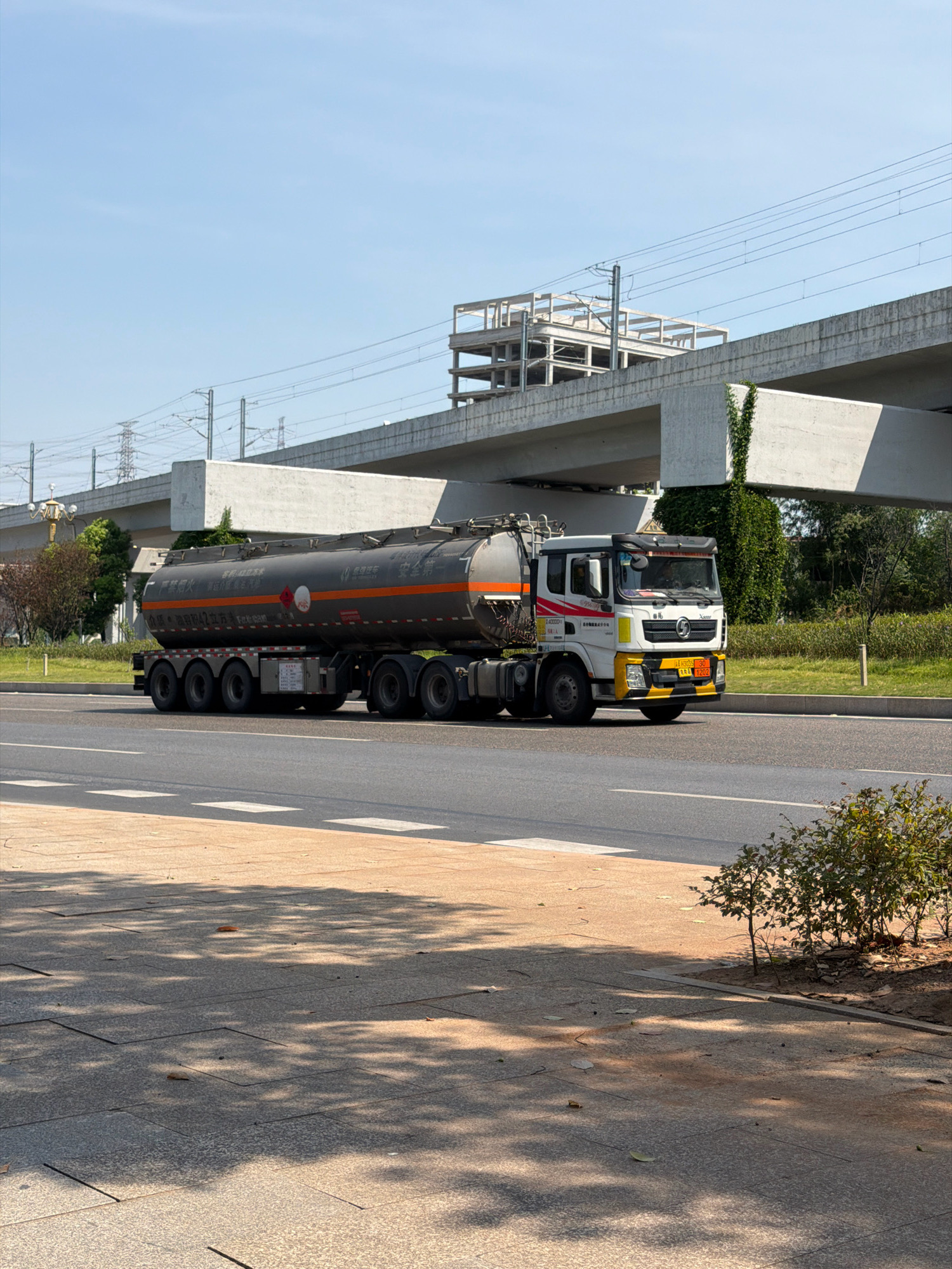 A tanker truck is driving on a road in Jiangjin, China. The truck is grey with orange stripes and has a long cylindrical tank. The truck is on a multi-lane road with a yellow dividing line. In the background, there is a concrete overpass with construction elements and electrical lines. There are also trees and grass on the side of the road. The sky is clear and blue. The foreground shows a paved walkway with dappled sunlight and shadows from trees.