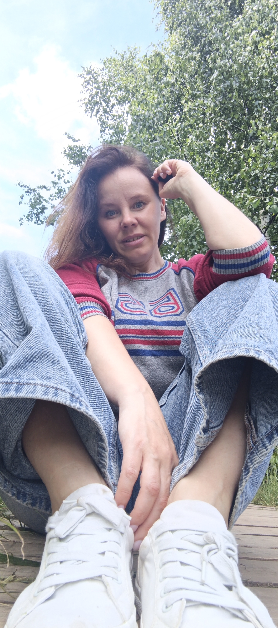 A Caucasian woman with shoulder-length brown hair is sitting on a wooden platform outdoors, with her legs spread and bent, wearing light blue denim jeans and white sneakers. She is looking at the camera with a slight smile, her right hand resting on her head. She is wearing a grey sweater with red and blue stripes on the sleeves and a red, blue, and white pattern on the chest. Behind her, green foliage from trees and a light blue sky with white clouds are visible. The image appears to be taken from a low angle, looking up at the woman. The location is a natural setting, possibly a park or forest, with no visible city structures.