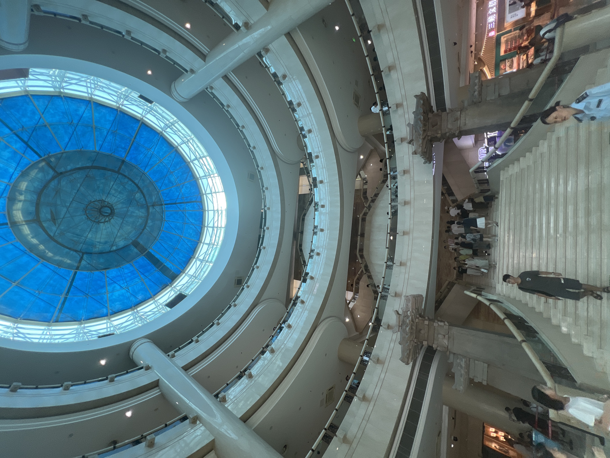 This is a wide-angle, interior shot of a multi-level shopping mall in Yuzhong District, China, with a focus on the central atrium and a large, blue, domed skylight. The architecture is modern, with curving balconies and large, cream-colored pillars supporting the structure. Numerous people are visible on different levels, some walking up a wide, central staircase, others standing on the balconies. The overall lighting is bright and airy, with the blue of the skylight dominating the upper portion of the image. Decorative stone pillars, reminiscent of ancient Chinese architecture, flank the staircase on the lower levels. Storefronts with signage are visible in the background.