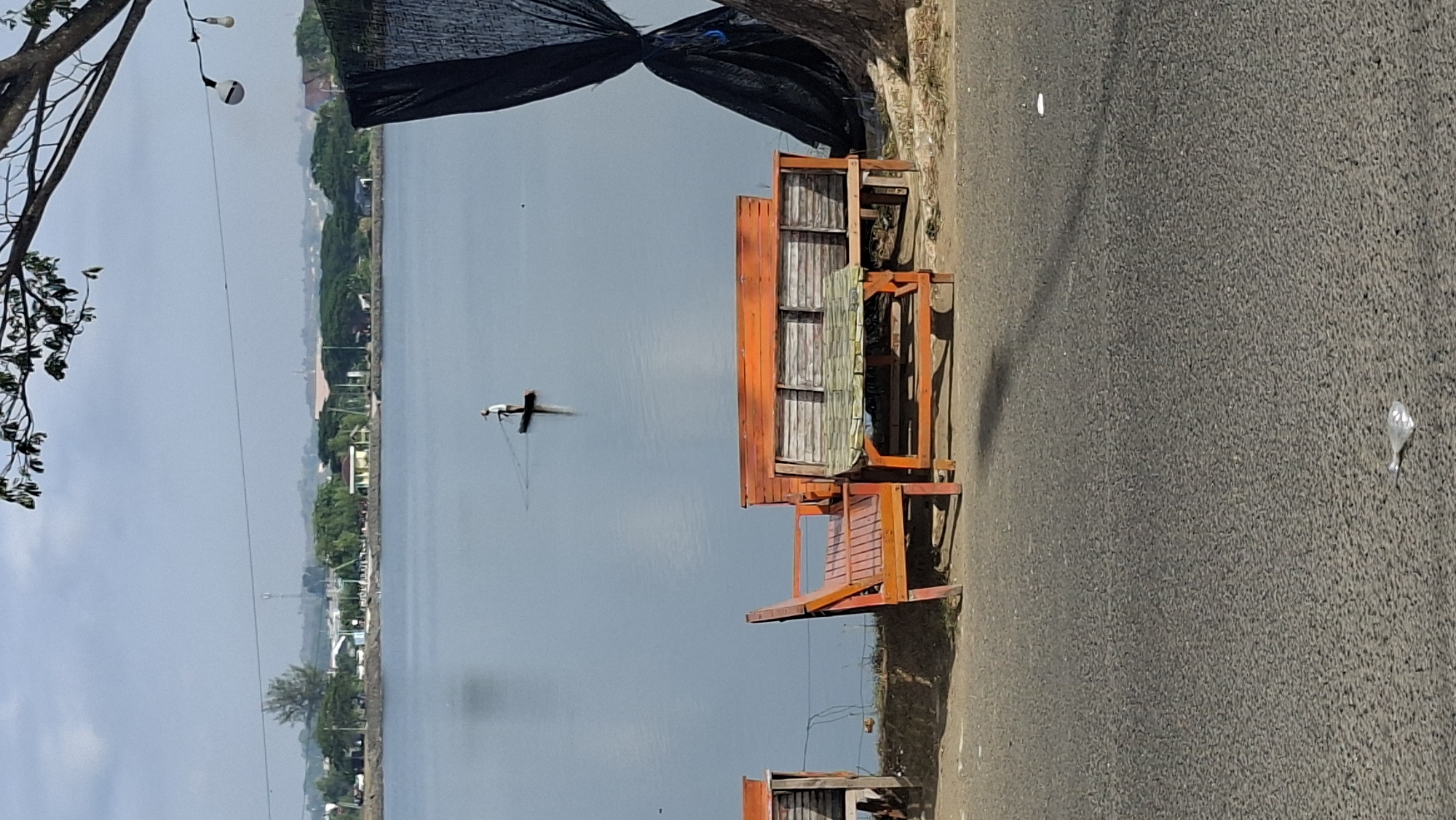 The image captures a serene view of the ocean at Lhokseumawe, Indonesia. In the distance, a lone individual stands on a small raft or boat, engaged in fishing. The water is calm and reflects the overcast sky. In the foreground, there are two wooden benches, one with a backrest and the other a long bench with a table. A large black tarp hangs to the right of the scene, partially obscuring a tree. The ground in the foreground is asphalt, with some scattered debris. The overall impression is one of a quiet, peaceful coastal area.