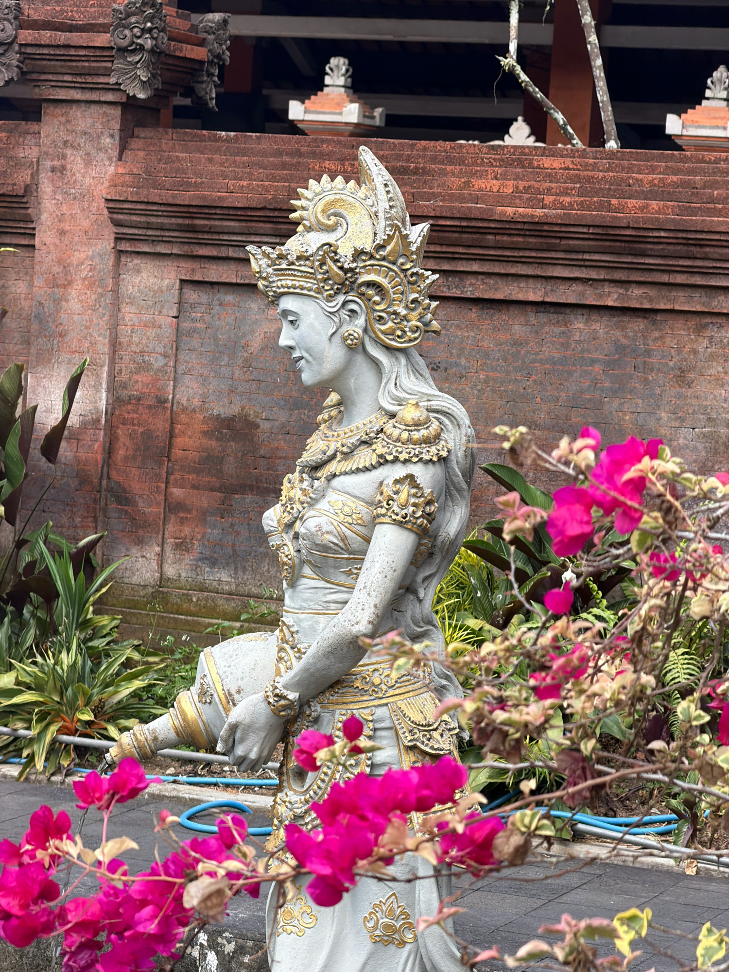 A statue of a woman, likely a deity or a dancer, stands in the foreground, rendered in a grey material with ornate gold detailing. She is depicted in profile, facing left, wearing an elaborate headdress, jewelry, and a detailed costume. Her hair is styled in long waves, and she appears to be holding a vessel from which water is flowing. The statue is positioned in front of a textured, reddish-brown brick wall that forms the background. Lush green foliage, including large-leafed plants and bright pink bougainvillea flowers, are prominent in the foreground and to the sides, partially obscuring the statue and its surroundings. The ground appears to be paved with stone tiles. The overall scene suggests a temple or a cultural site. Based on the architectural style of the brickwork and the statue's ornamentation, this image likely depicts a scene from Tampaksiring, Indonesia, possibly within the grounds of a temple complex such as Tirta Empul or Gunung Kawi, which are known for their ancient sanctuaries and water features.