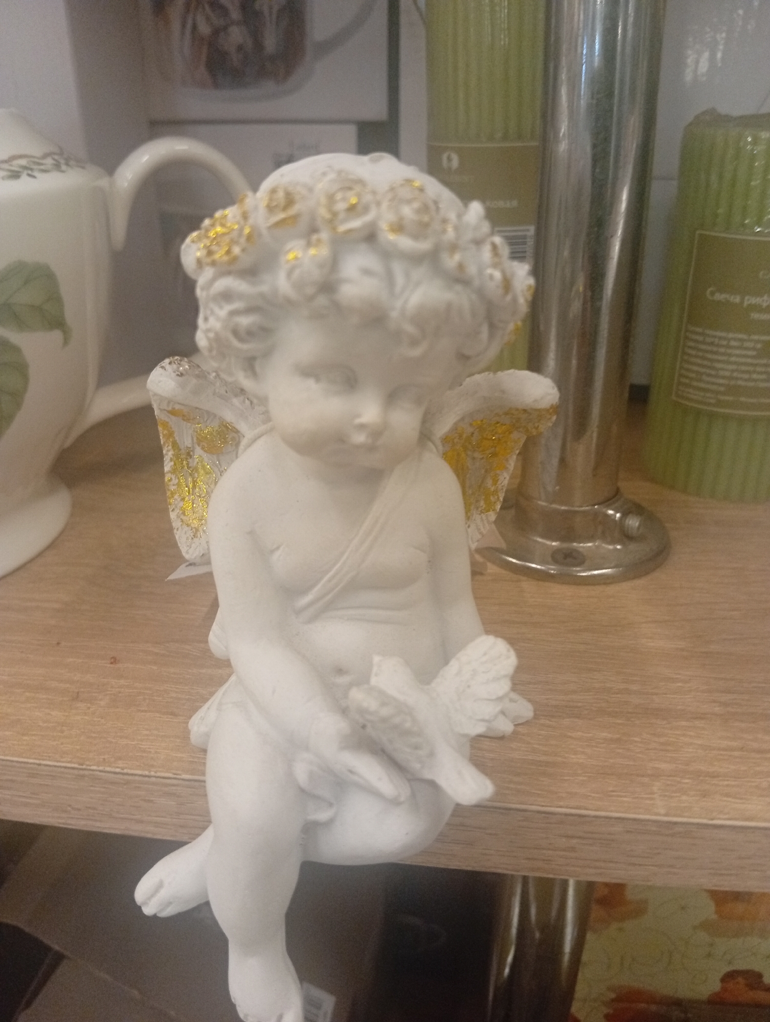 A white ceramic figurine of a cherub sits on a light brown wooden shelf. The cherub has a wreath of golden flowers on its head and golden accents on its wings and tunic. It is holding a small bird in its hands. Behind the cherub are several tall green candles and a silver cylindrical object. The background is out of focus, but appears to be a shelf with more items. The scene is indoors and appears to be a decorative display. The specific location in Pushkino, Russia is not determinable from the image alone.