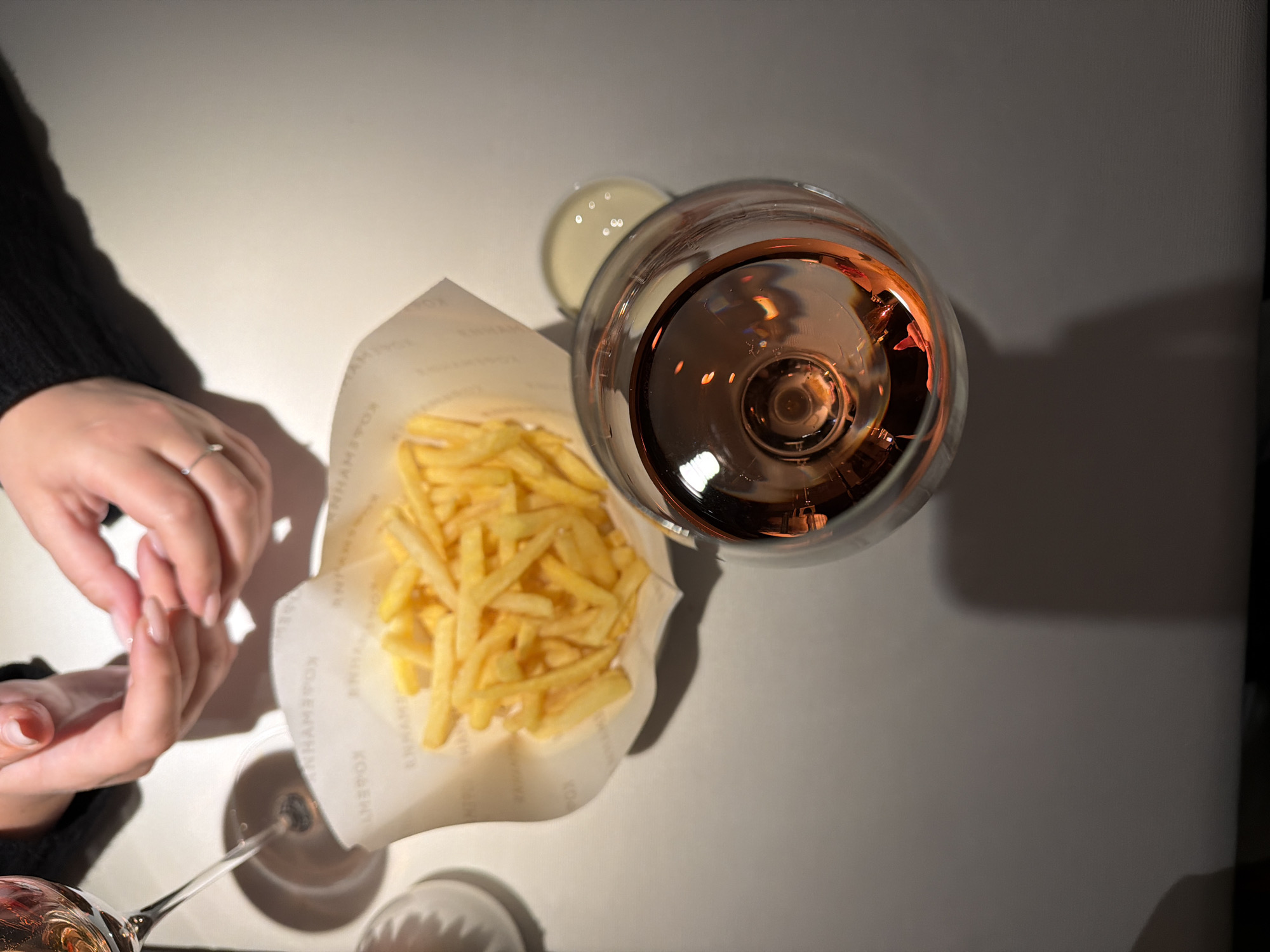 A top-down shot shows a pair of hands resting next to a paper cone filled with french fries. Beside the fries is a small bowl of dipping sauce and a wine glass filled with a rose-colored liquid. The image is taken indoors, on a table surface. The environment appears to be a restaurant. The city and state is Unknown, Unknown.