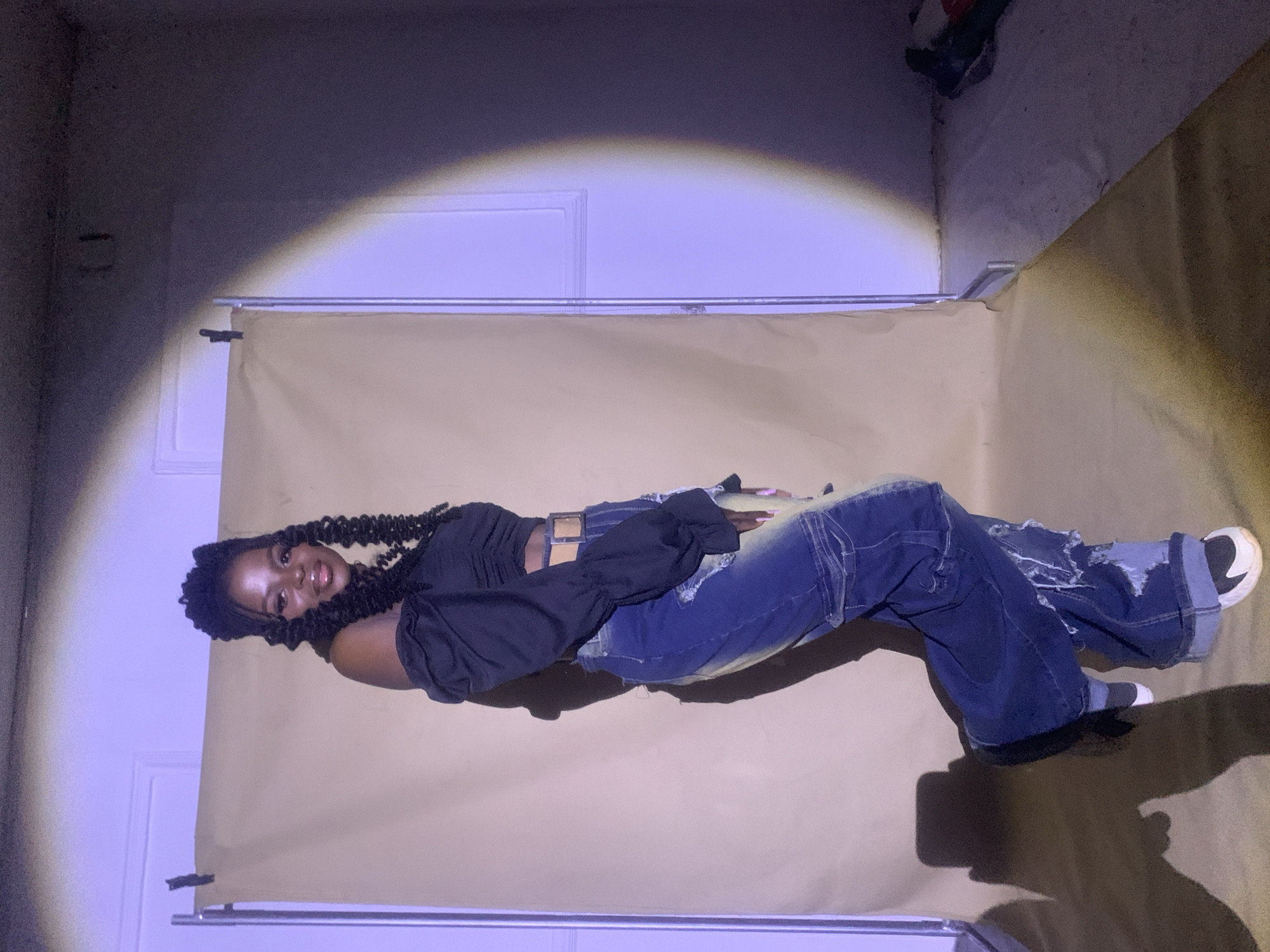 A young Black woman is reclining against a neutral-toned backdrop that is hung on a frame. She wears a black, long-sleeved crop top, baggy distressed jeans, and white sneakers. The backdrop is set up in a room with white walls. There is a door behind the backdrop. The lighting is uneven. The location is not discernible as Jos, Nigeria, from the visual information provided.