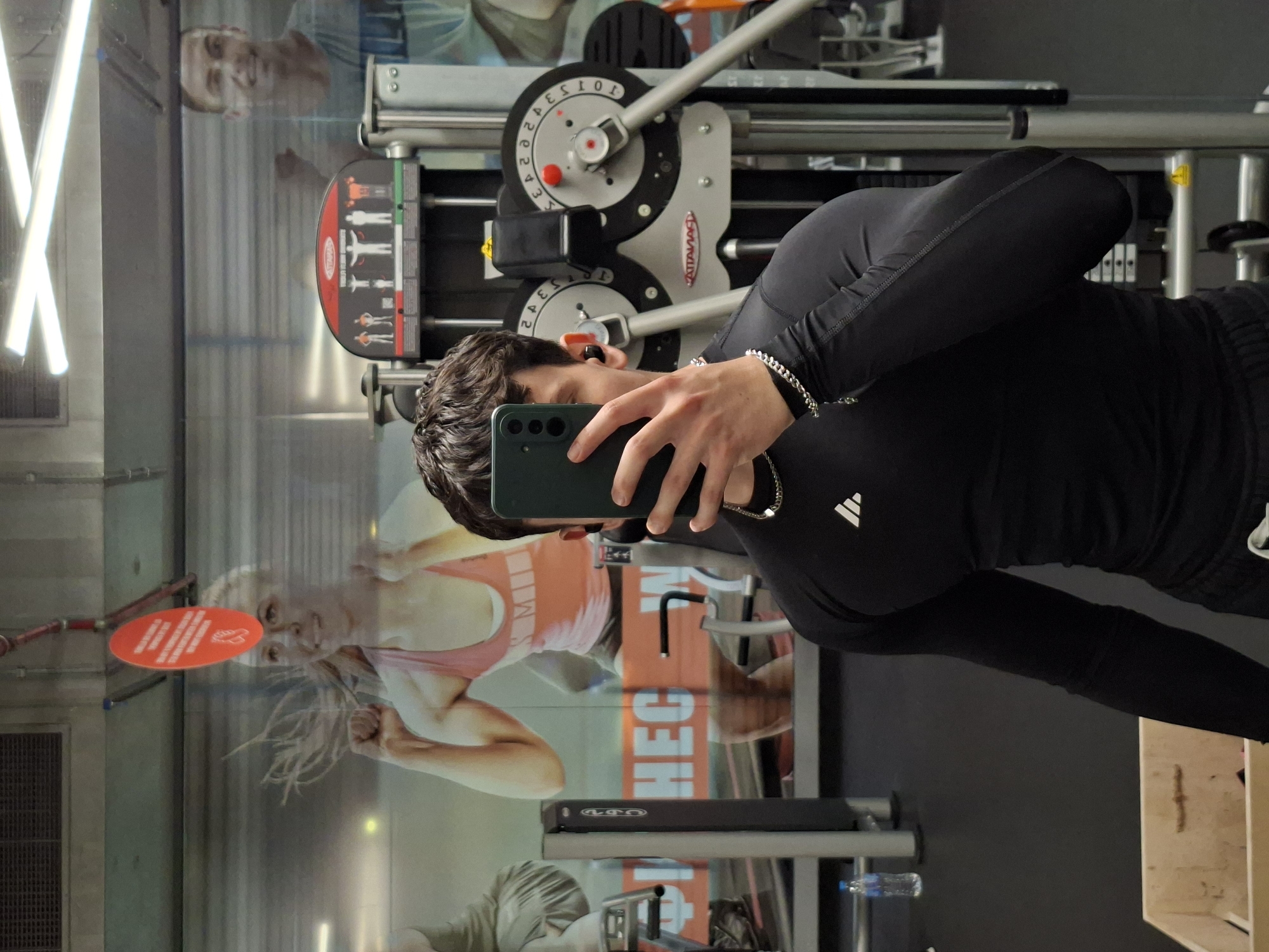 A young man is taking a mirror selfie in a gym. He is wearing a black long-sleeved shirt and black shorts, and he has earbuds in his ears. He is holding a smartphone in his right hand. In the background, there are gym machines and posters of people exercising. The overall lighting is dim, typical of a gym environment. The specific location of the gym in Plovdiv, Bulgaria, cannot be determined from the visual information provided in the image.