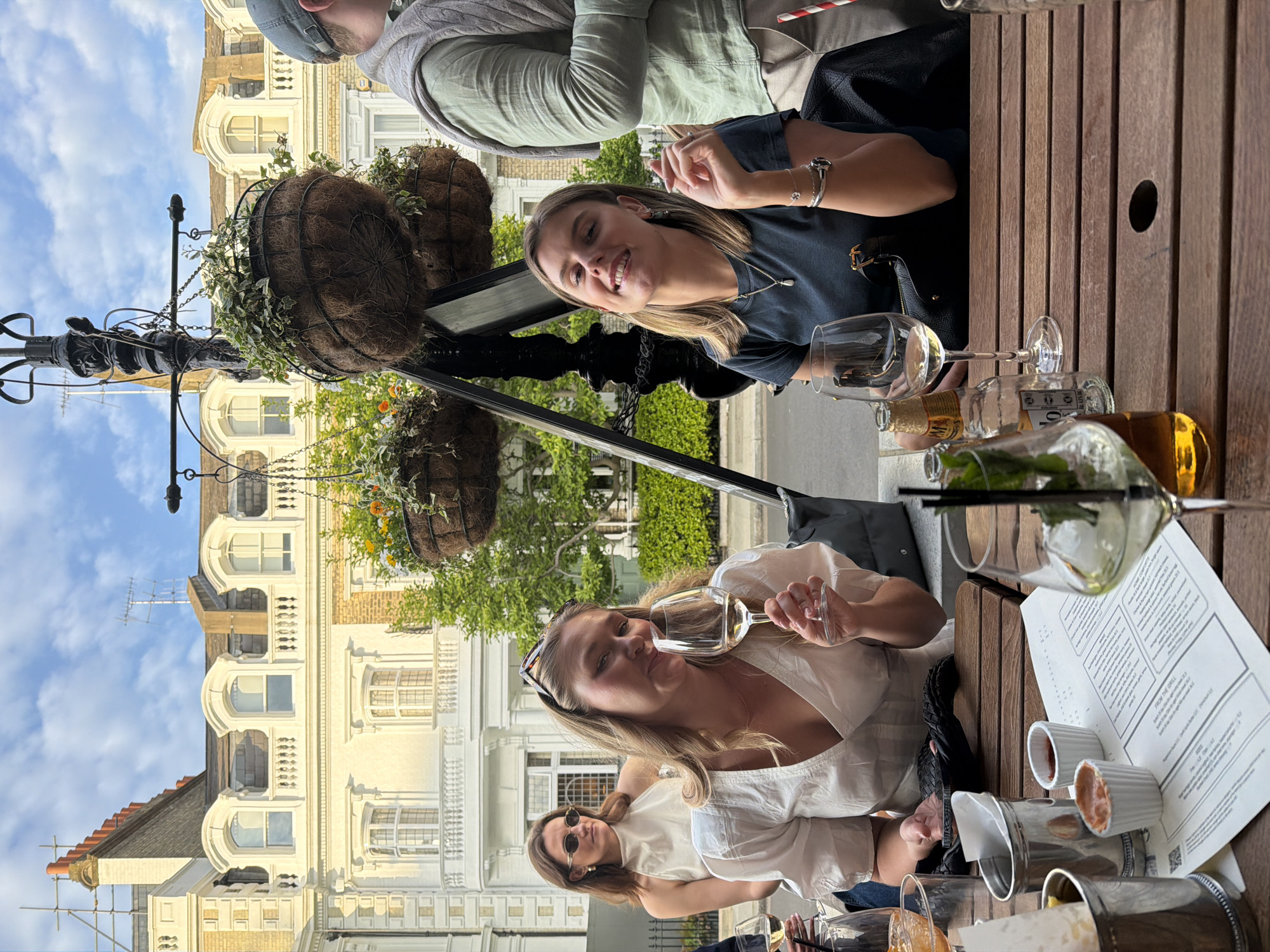 This image captures an outdoor dining or socializing scene in London, featuring several individuals enjoying drinks and conversation in bright daylight. The weather appears pleasant, with a clear blue sky and some white clouds.
In the foreground, several people are seated around wooden slatted tables. Prominently featured are four women. One woman with blonde hair, wearing a white v-neck top and sunglasses on her head, holds a wine glass to her mouth, smiling subtly. Behind her, another woman with sunglasses is also looking towards the camera. At an adjacent table, a different woman with blonde hair, wearing a dark blue t-shirt, smiles broadly at the camera. She wears a necklace and a bracelet. Partially visible to her right is a man in a green long-sleeved shirt and a grey cap.
The tables are set with wine glasses, some containing white wine, and various bottles, including clear and amber ones, and a partially visible golden-foiled bottle. Small white ramekins, possibly holding sauces or dips, and a metal container are also present. Paper menus are visible on the tables, with the heading "FROM THE GRILL" clearly legible.
The background showcases impressive European architecture, with multi-story buildings featuring ornate white facades, arched windows, and balconies, typical of London's urban landscape. A black lamppost adorned with hanging baskets filled with ivy and other greenery stands in the mid-ground. Scaffolding is visible on a building in the distance, and a rooftop antenna is silhouetted against the sky. The overall scene suggests a relaxed and social gathering, with participants appearing happy and engaged.
