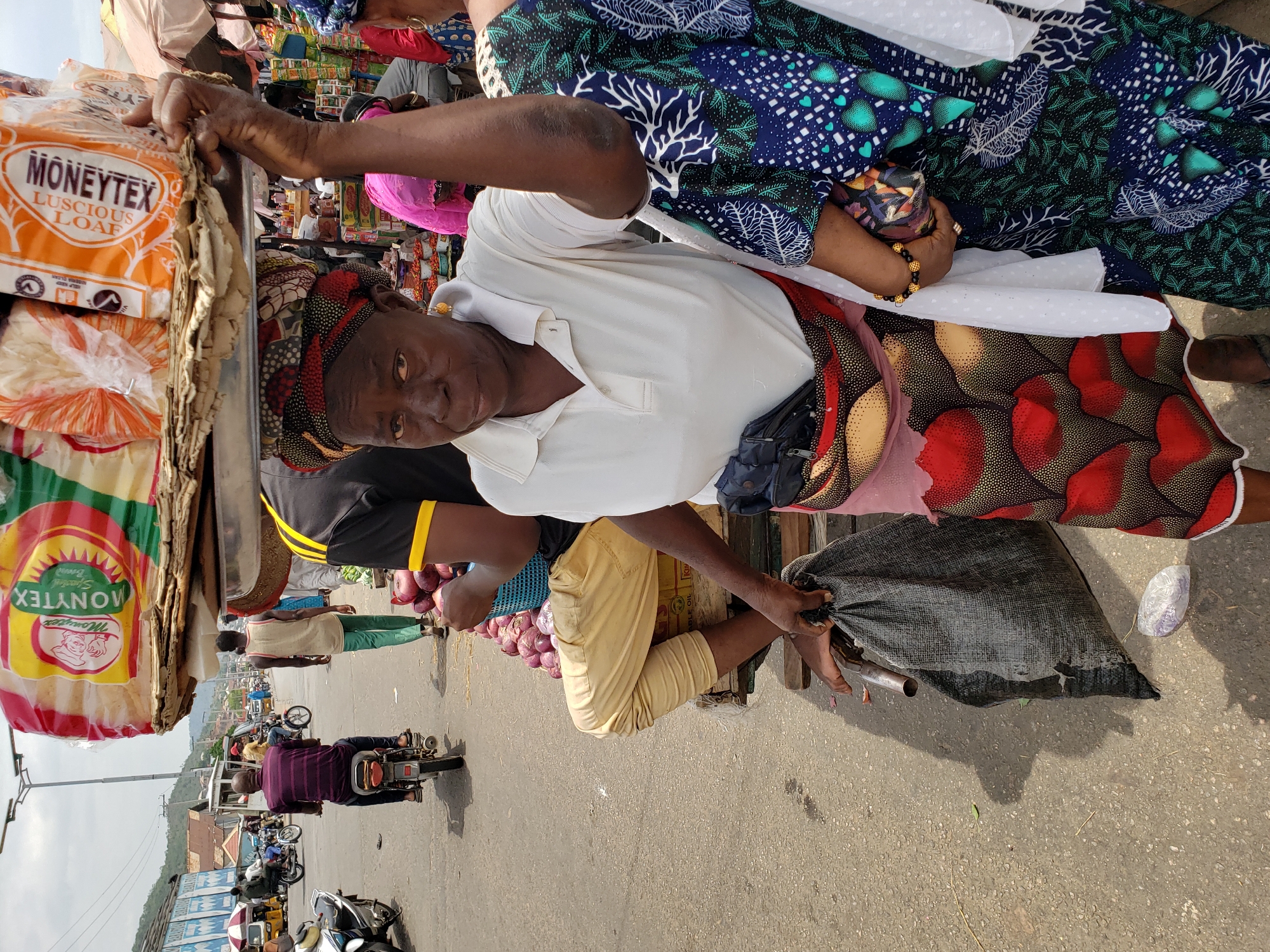 An overhead, medium-angle, eye-level, daytime, outdoor shot shows a woman in Lokoja, Nigeria, seated in front of a cart holding bread.
The woman is an adult with dark skin and short black hair. She wears a colorful headscarf, a white collared shirt, and a patterned skirt with red and brown colors. She has a black bag hanging from her shoulder. She is looking toward the viewer, and she has her left hand on a stack of bread. In front of her is a black plastic bag.
To the left of the woman, there is a cart holding loaves of bread. The bread loaves are labeled "MONYTEX LUSCIOUS LOAF" in orange, white, green, and red packaging. Behind the woman, there is another individual with onions.
In the background of the image, there is a busy street with motorcycles and other vehicles driving by. There are also buildings and trees visible. The road appears to be asphalt. The sky is pale and lightly cloudy.
