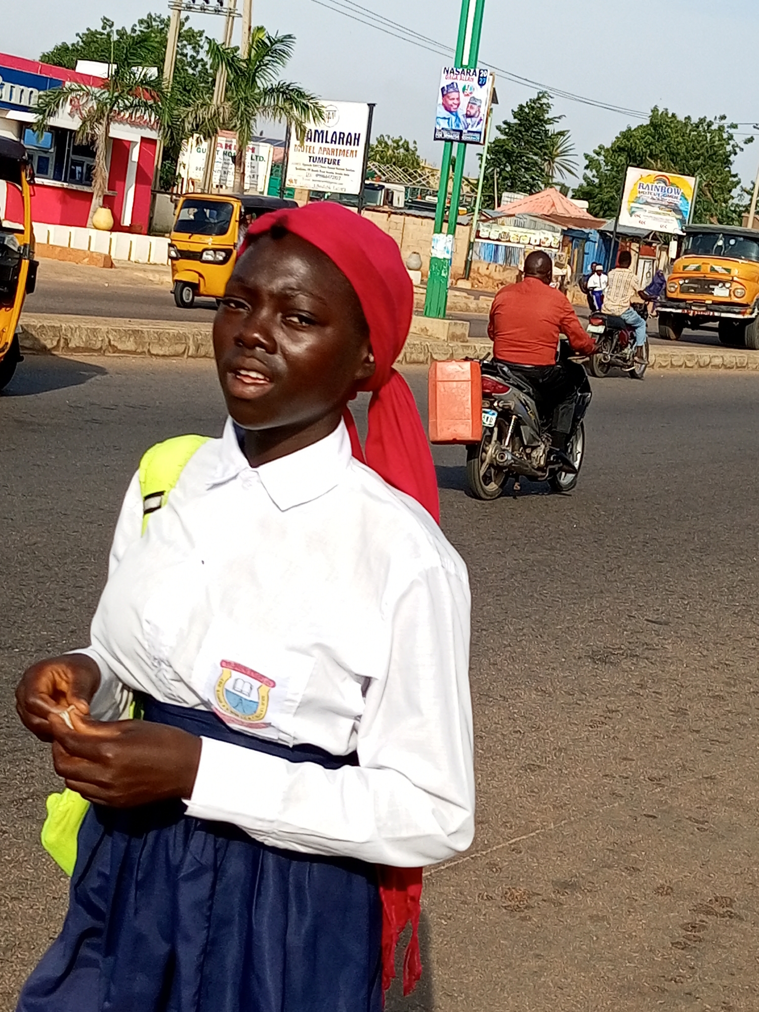 A young Black female, wearing a white collared shirt and a navy blue skirt, stands on an asphalt road. Her hair is covered with a red headscarf. She carries a bright yellow backpack on her left shoulder. Her right hand is raised, and she appears to be holding a small white object. The background shows a street scene in Tempure, Nigeria, with traffic, buildings, and vegetation. Various vehicles are visible, including a yellow autocar, a yellow truck, and motorcycles. Billboards and street signs are also present. The lighting suggests it is daytime.